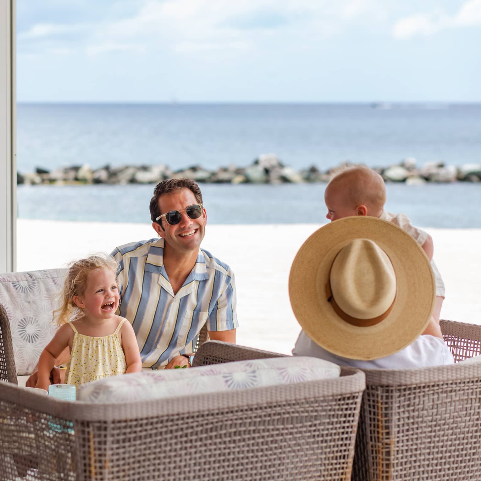 Three adults and two young children sit together facing each other on four rattan arm chairs with a beach in the background