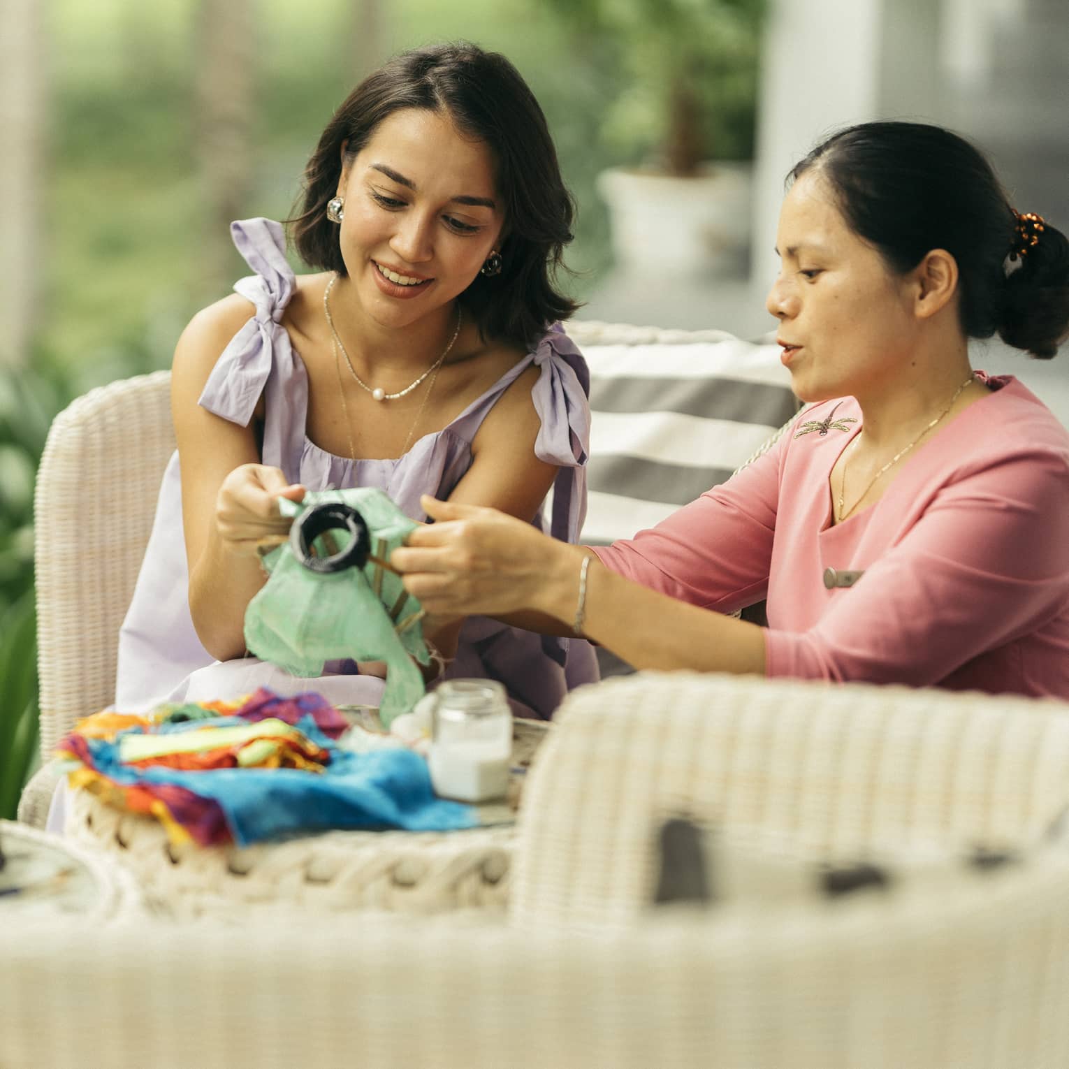 Two people engaged in a traditional craft activity, working together on fabric art while seated on a wicker patio set in a lush, green outdoor setting.