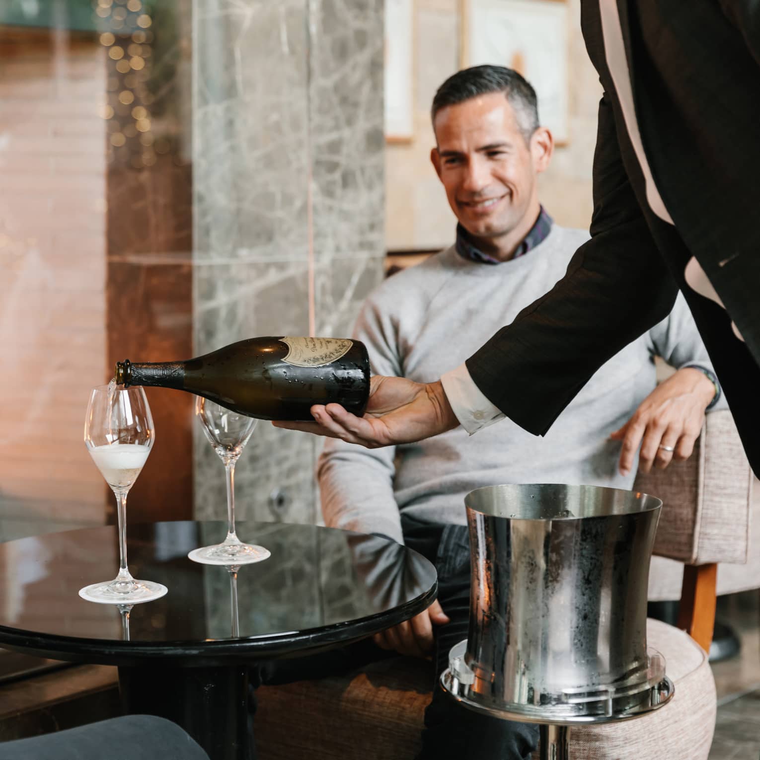 Two men being poured a glass of wine while sitting next to a fire.