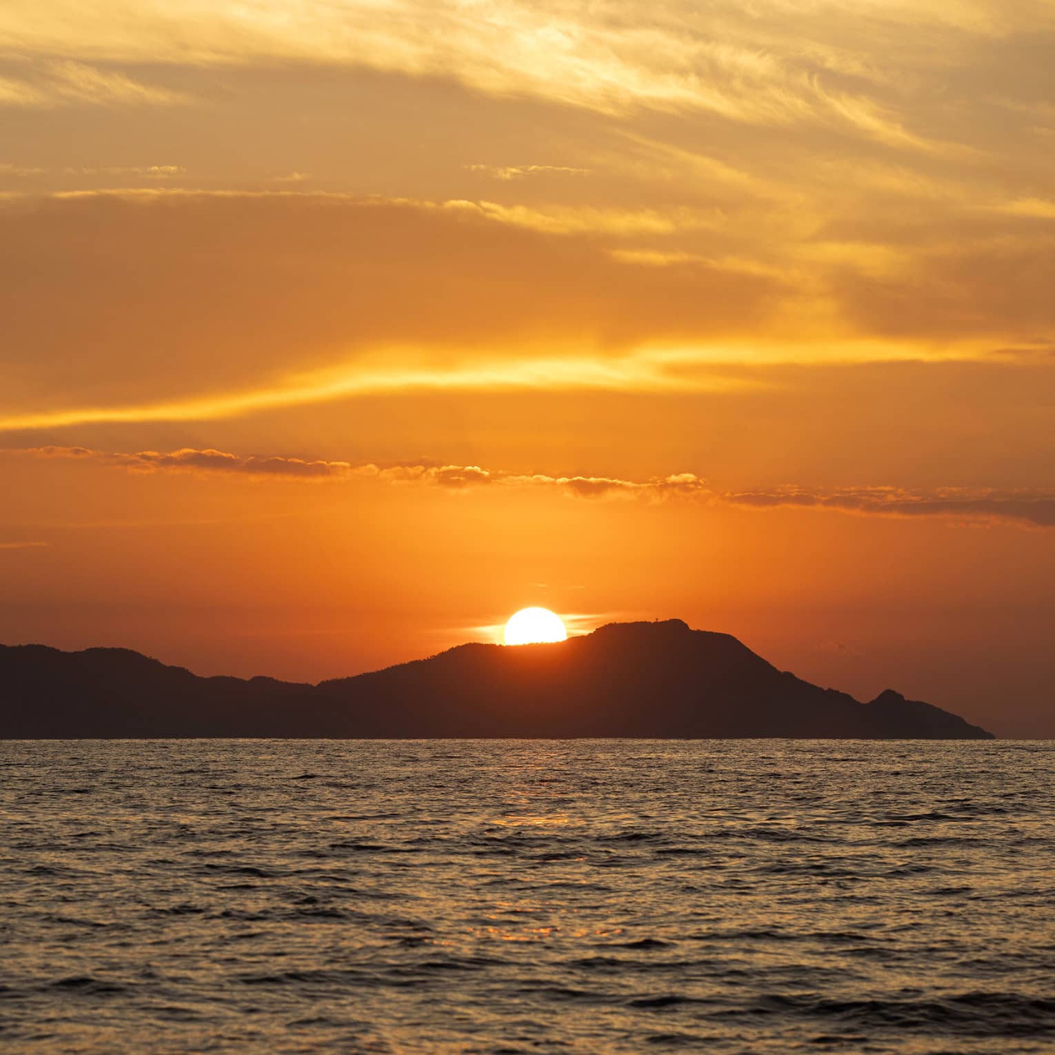 Beyond gently rippling waves, the blazing sun sets behind a hilly island, wispy clouds in the sky awash in its orange glow.