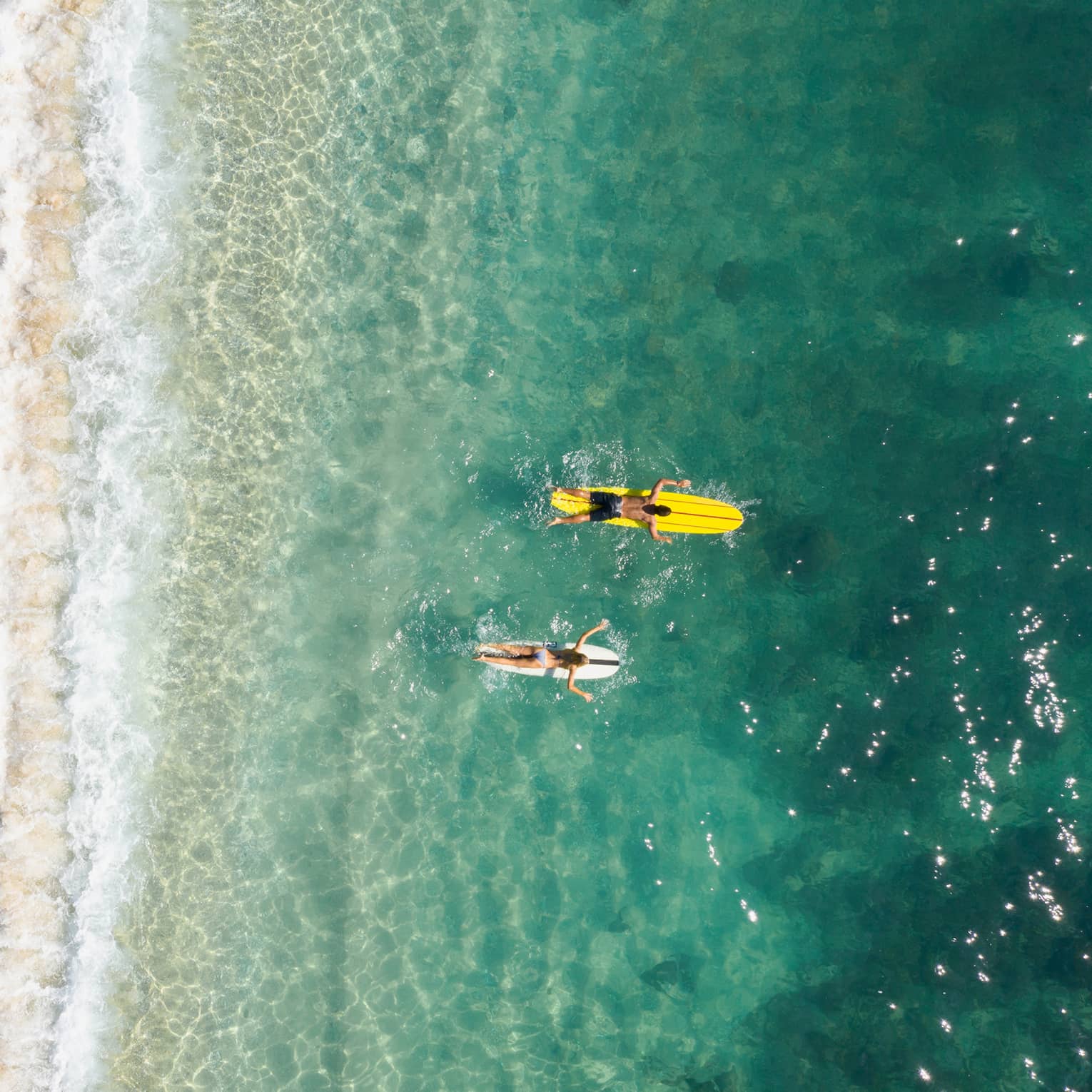 Bird’s-eye view of two people paddling their boards on their stomachs as frothy waves break on the beach behind them.