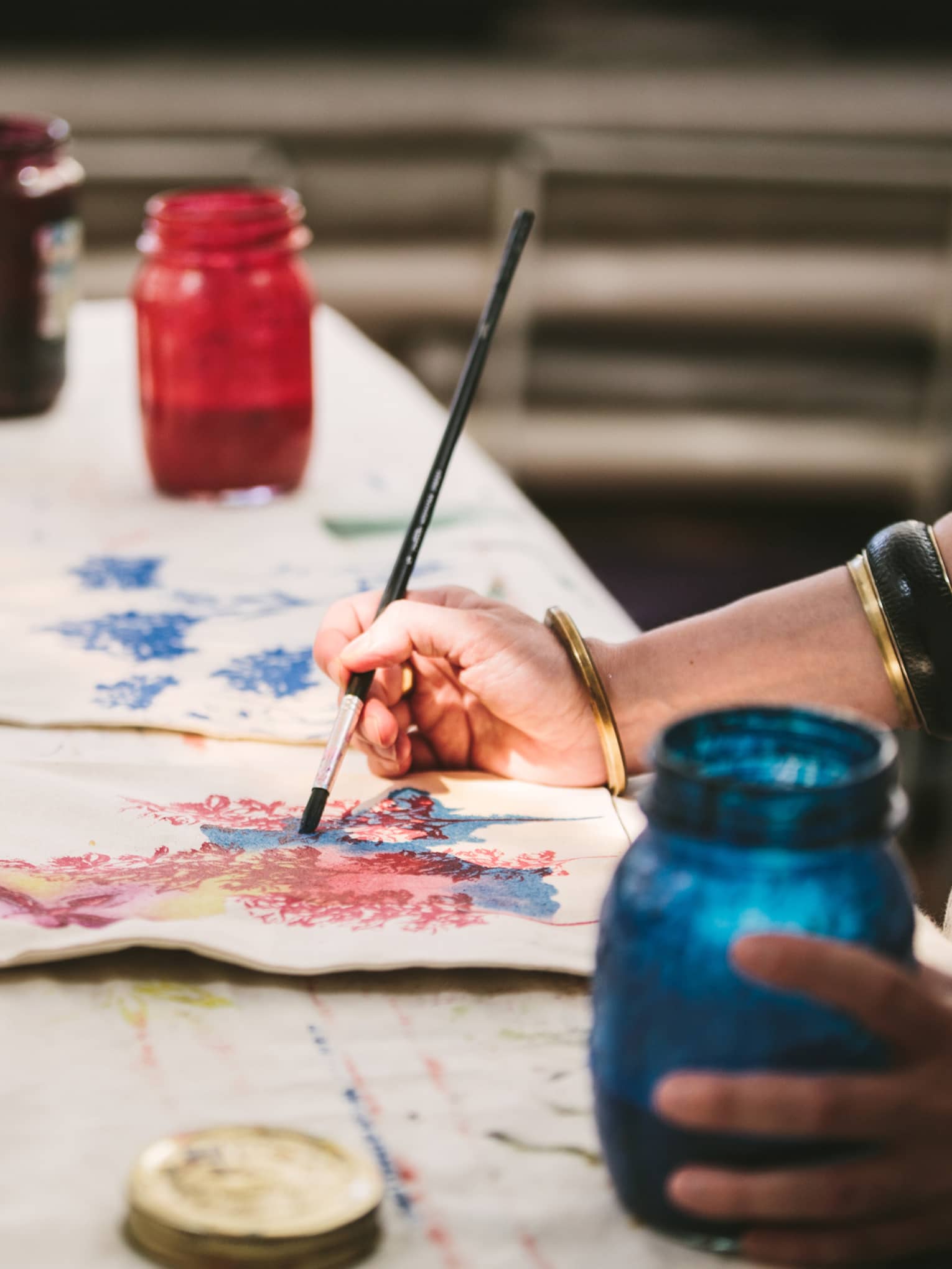 An artist using mason jars filled with red, blue, yellow and black paint to paint images of flowers on canvas tote bags.