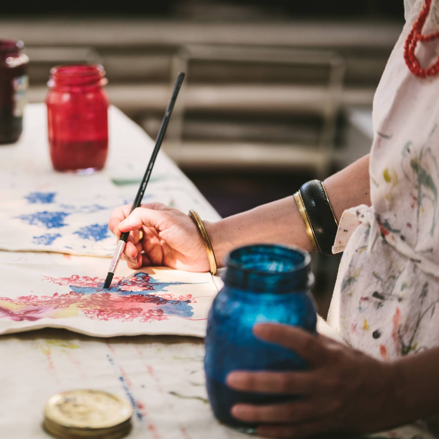 An artist using mason jars filled with red, blue, yellow and black paint to paint images of flowers on canvas tote bags.