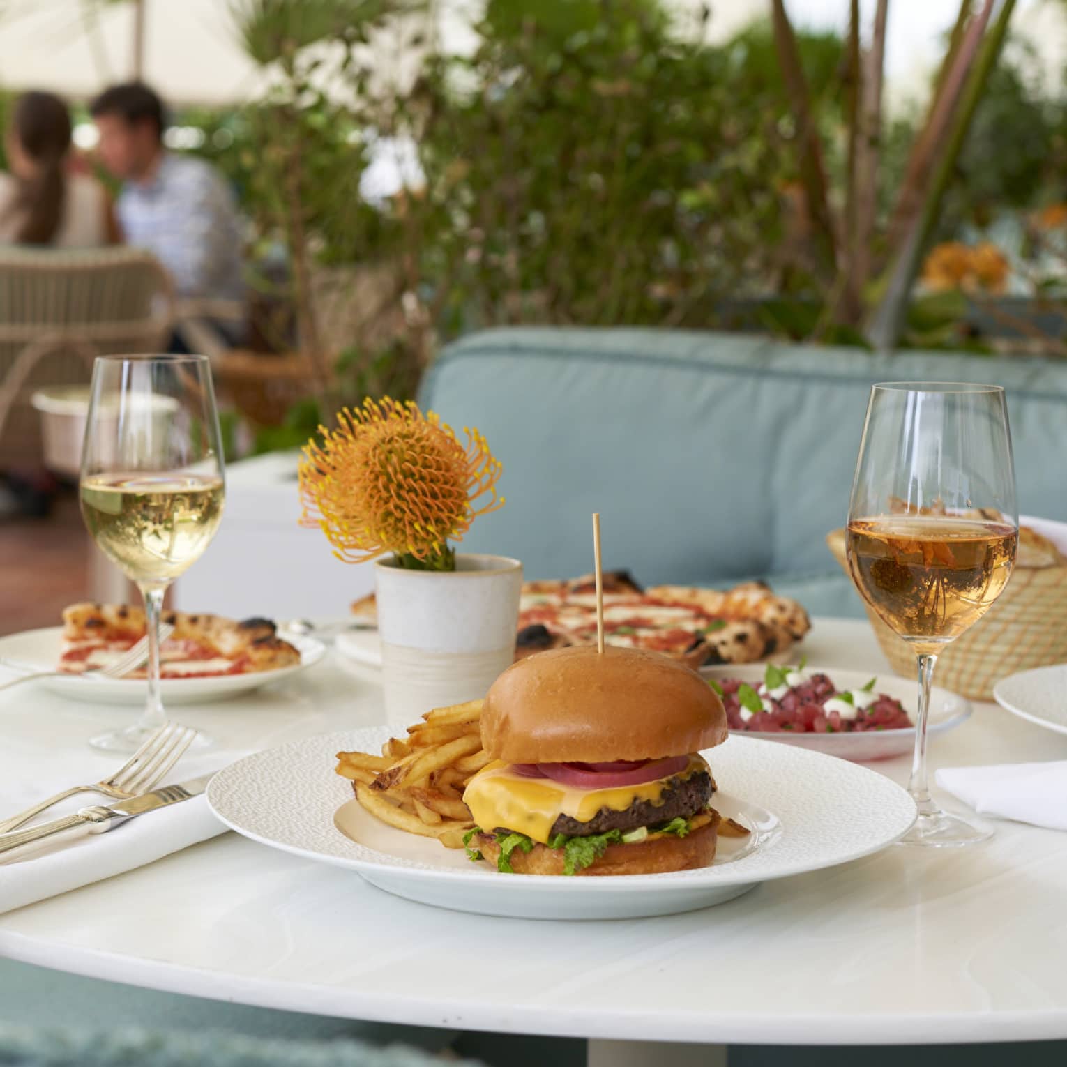 An outdoor dining table surrounded by chairs with plush blue cushions, topped with plates containing a cheeseburger, fries and pizzas and two glasses of wine