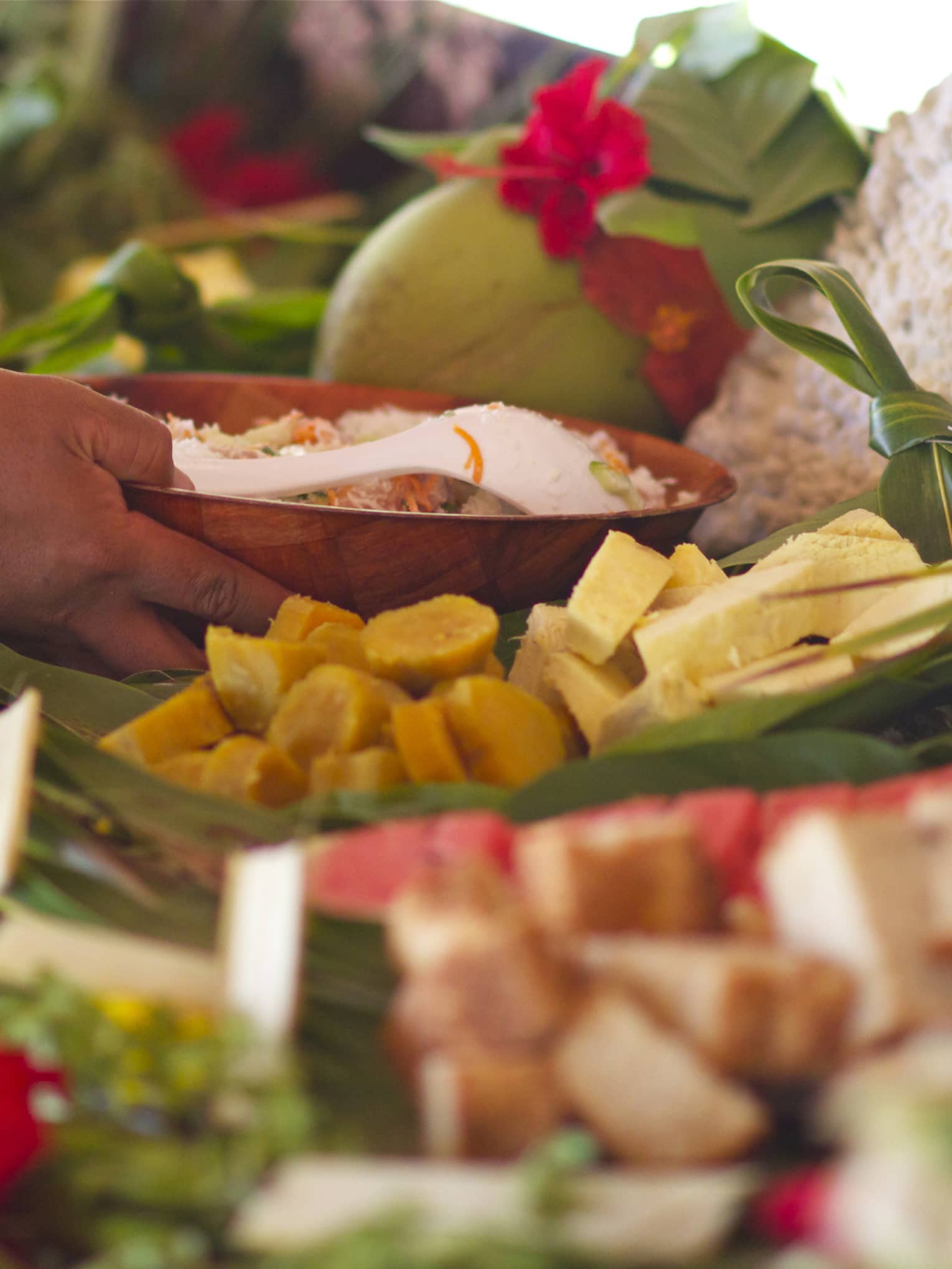 Hand grasps wooden bowl in display of fresh tropical fruit adorned with red flowers