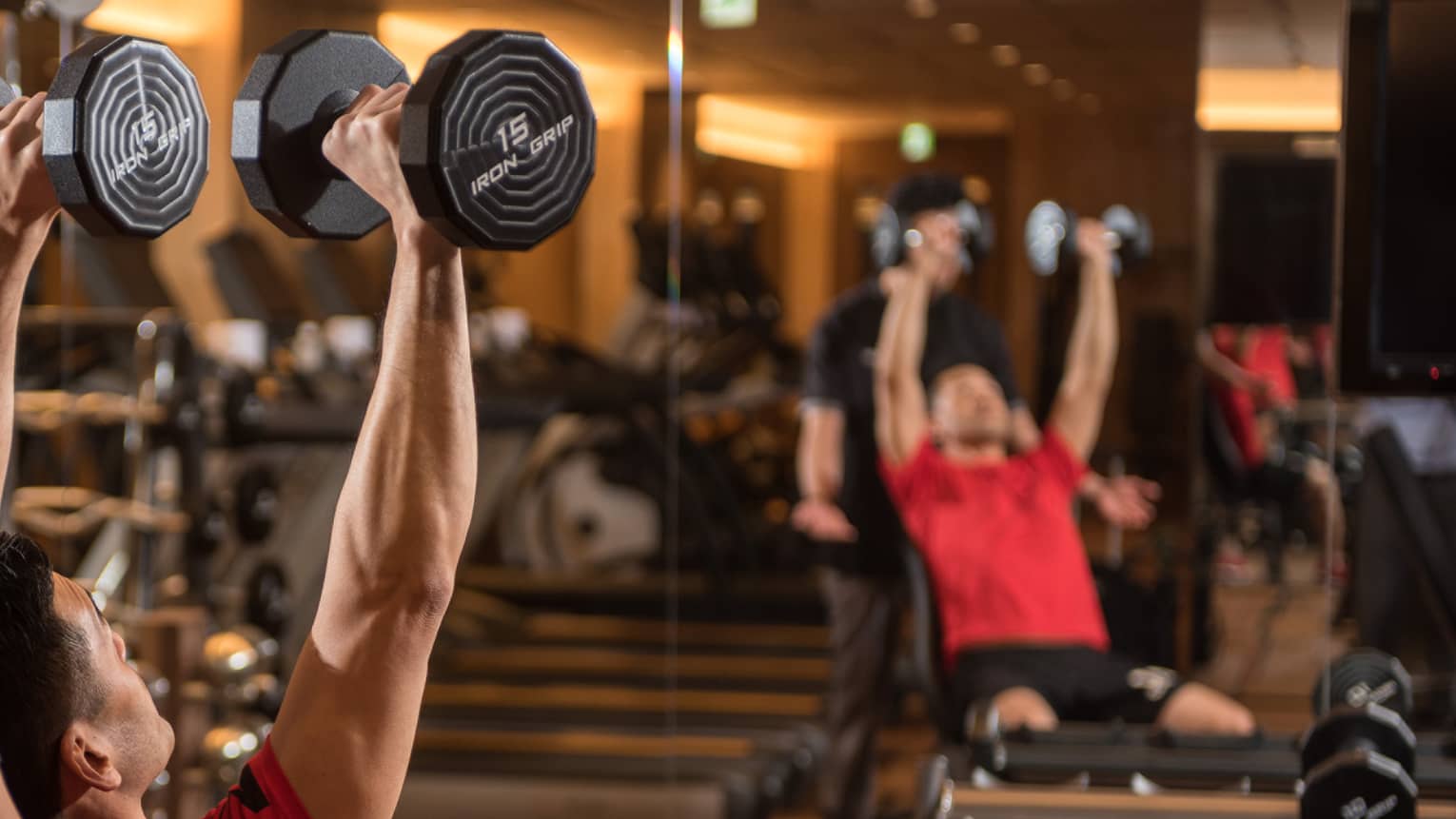 Man in chair in front of mirror lifts two 15 lb. weights above his head while personal trainer assists