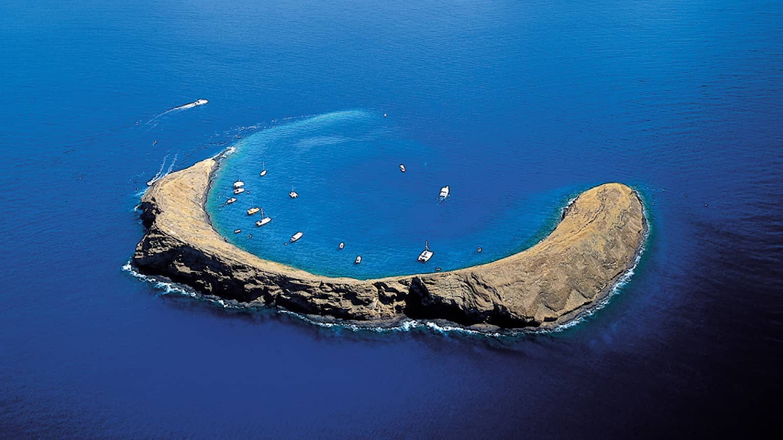 Aerial view of Molokini Crater, a crescent-shaped volcanic islet surrounded by clear blue ocean waters, with boats anchored nearby