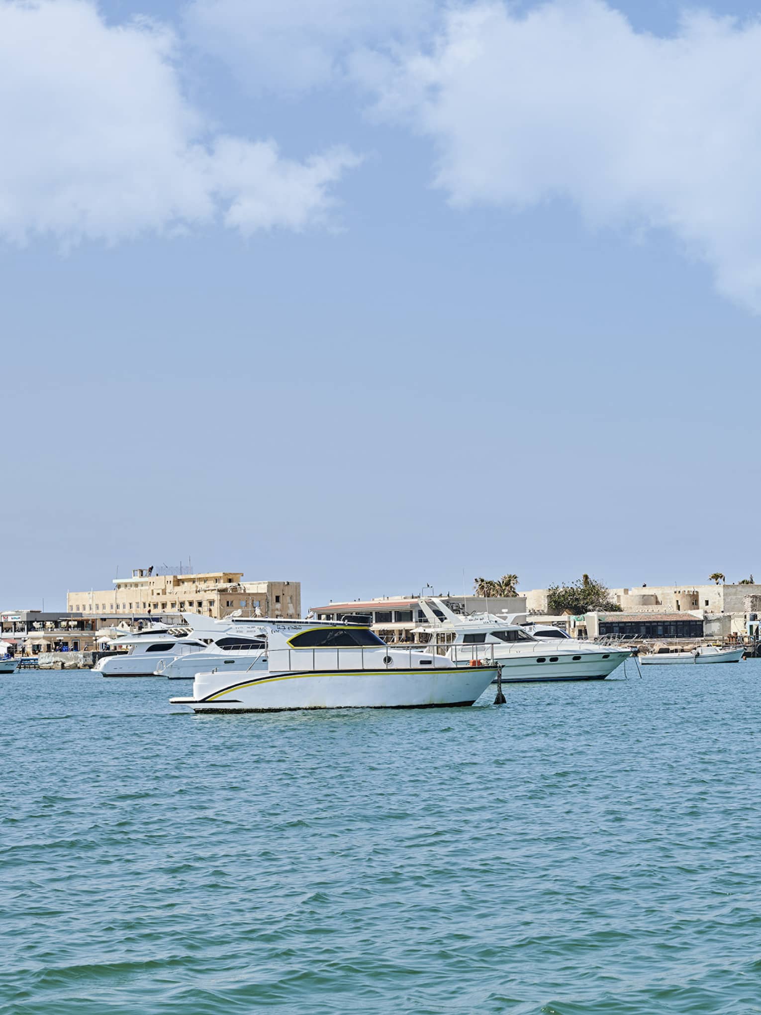 A distant sand-coloured fortress perched on a quay along a shore dotted with small yachts and fishing boats over calm waters.