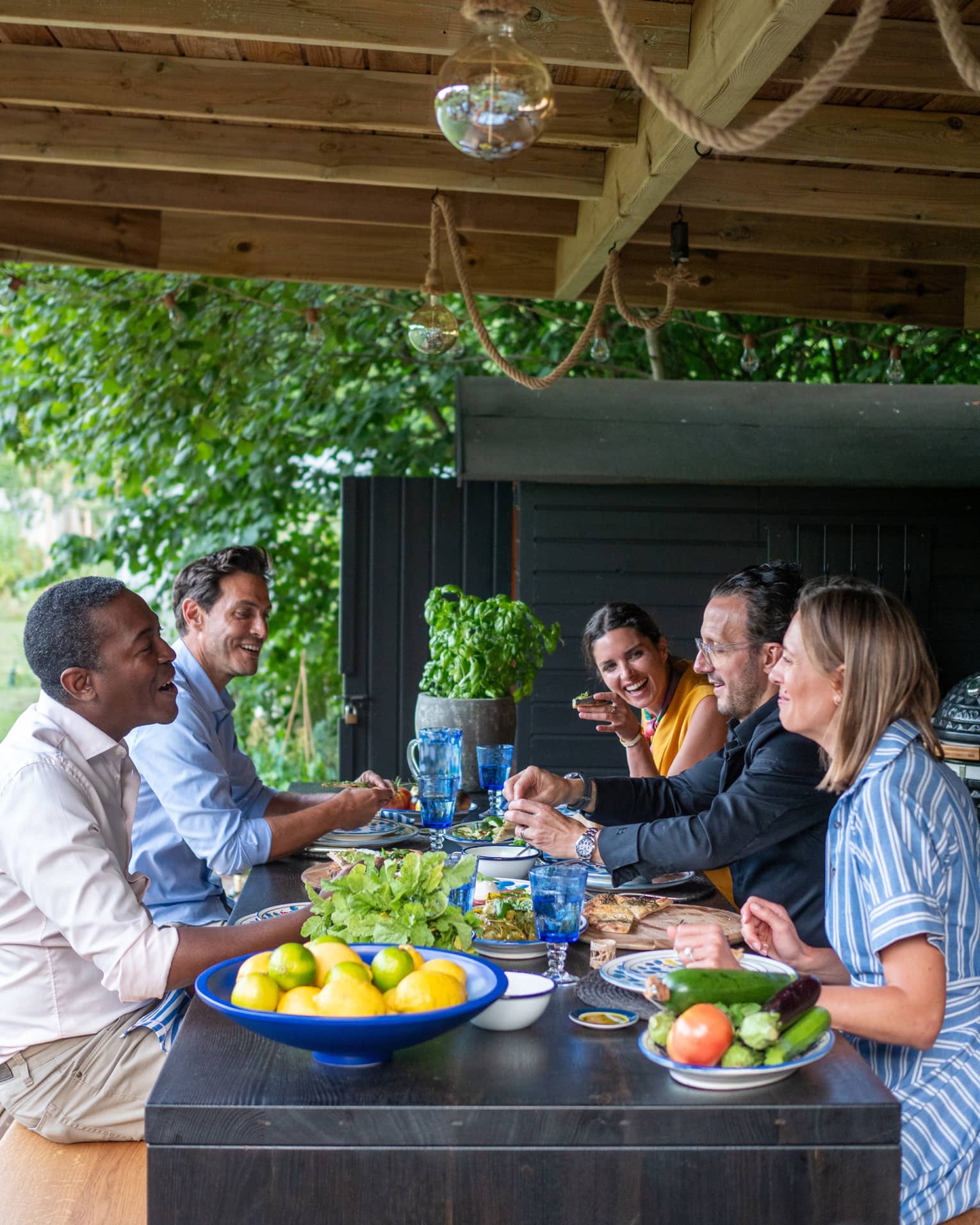 A smiling group of five people sit at a wooden table covered in dishes of flatbread, fresh vegetables and citrus.