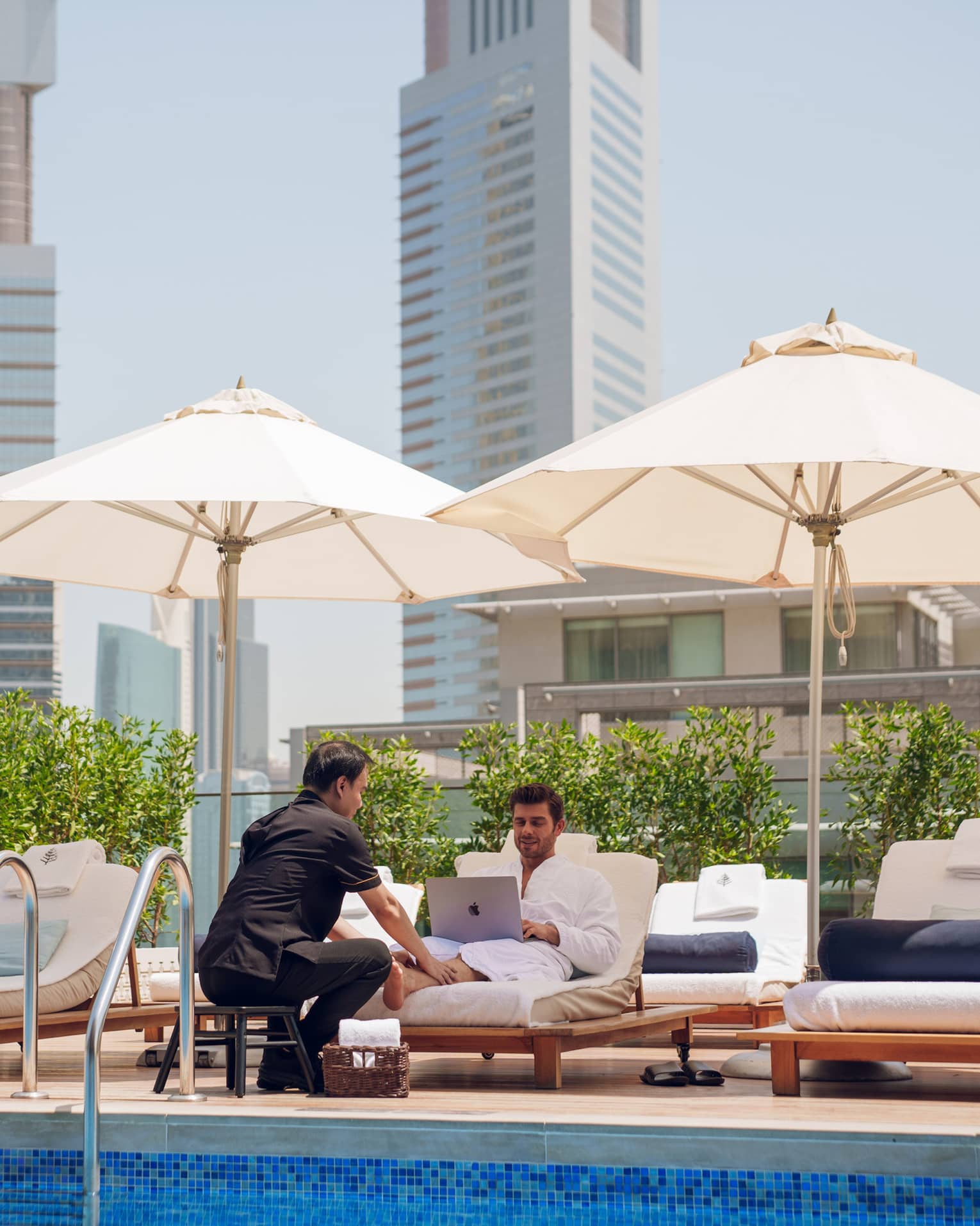 Guest sits on a poolside lounge chair looking at his laptop while a spa attendant rubs his feet