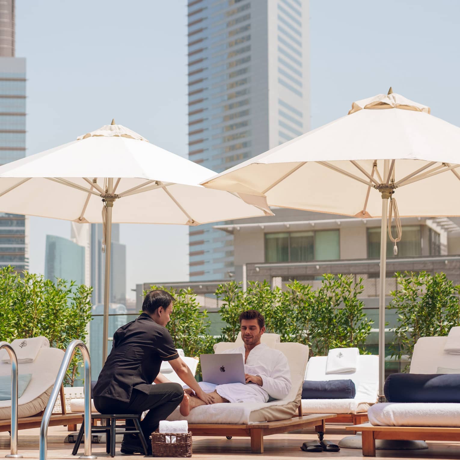 Guest sits on a poolside lounge chair looking at his laptop while a spa attendant rubs his feet