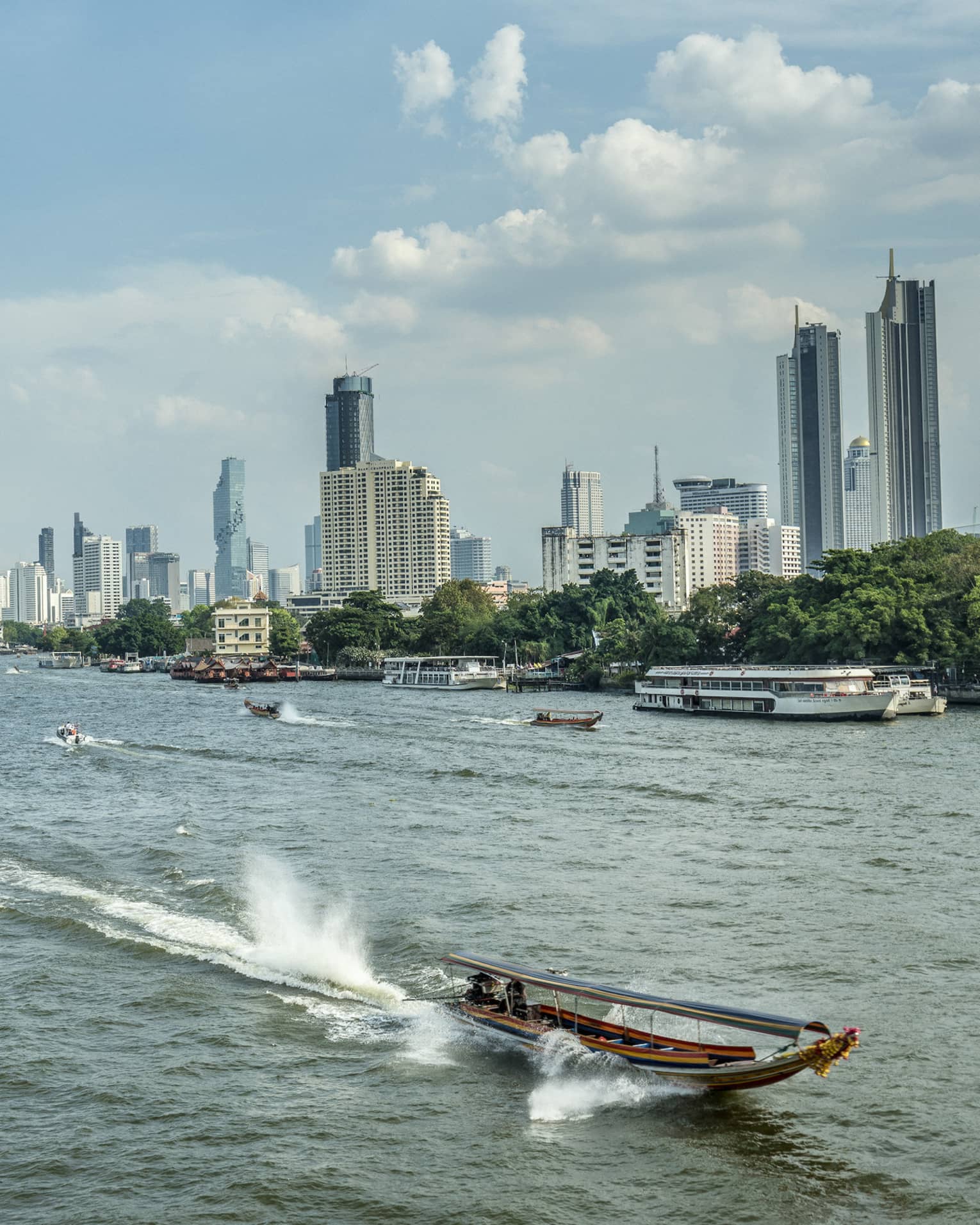 Longtail boat heading into foreground away from Bangkok skyline, leaving trail behind and surrounded by tall, splashing wake.