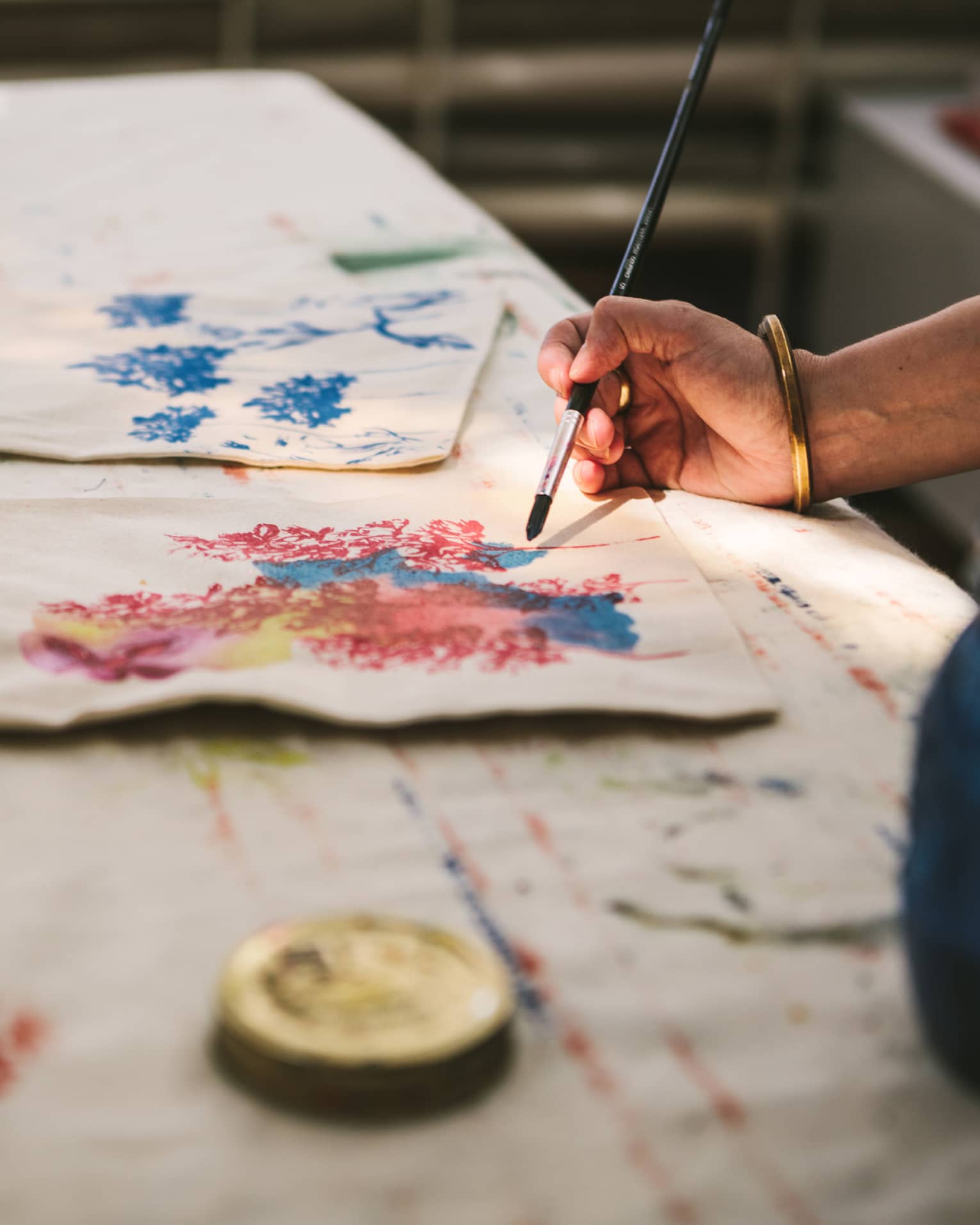 Close-up of a gold-bangled hand wielding a long brush to paint an intricate pattern on a cloth tote bag.