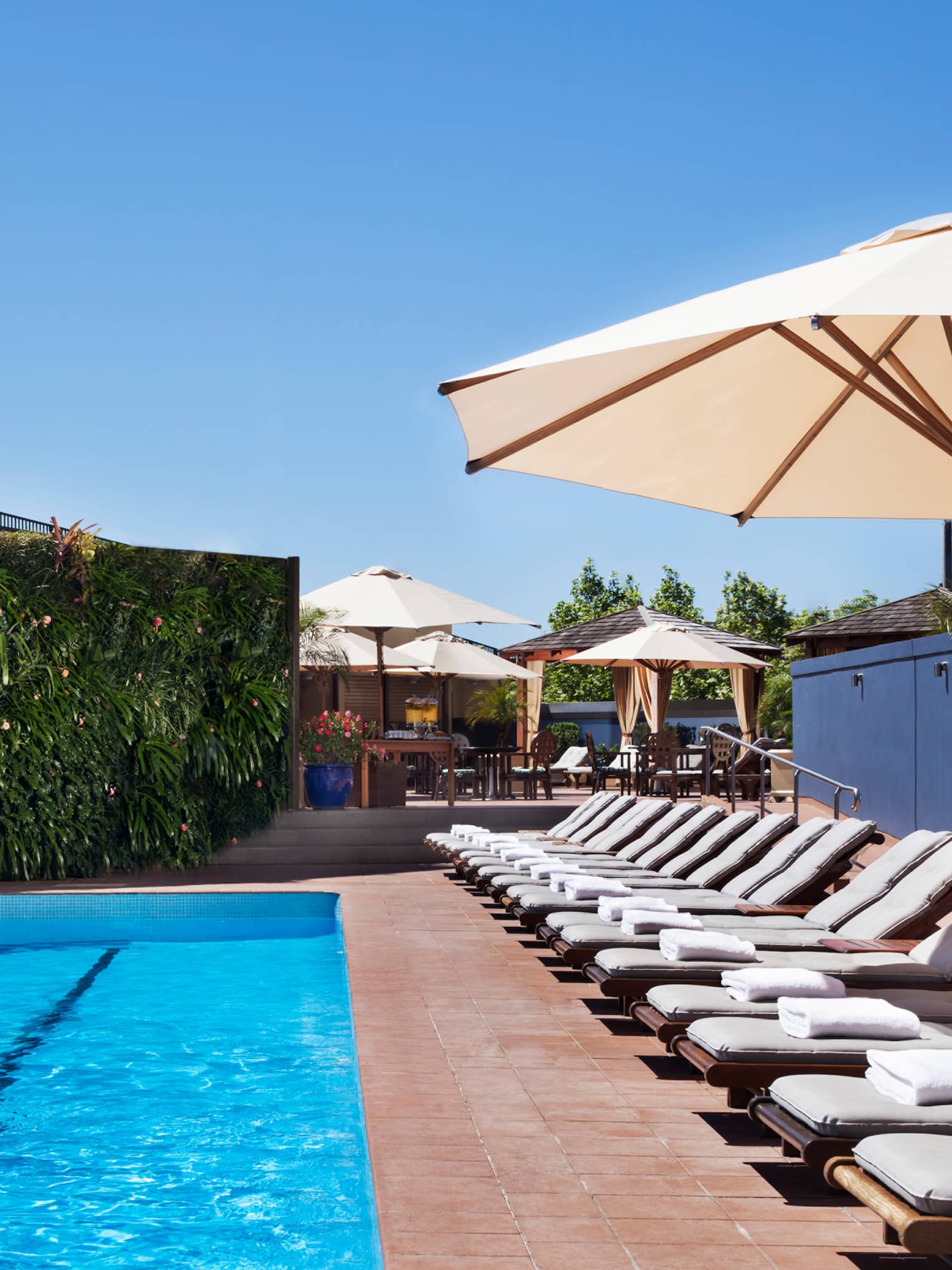 White lounge chairs lined up on deck under patio umbrella by blue outdoor swimming pool