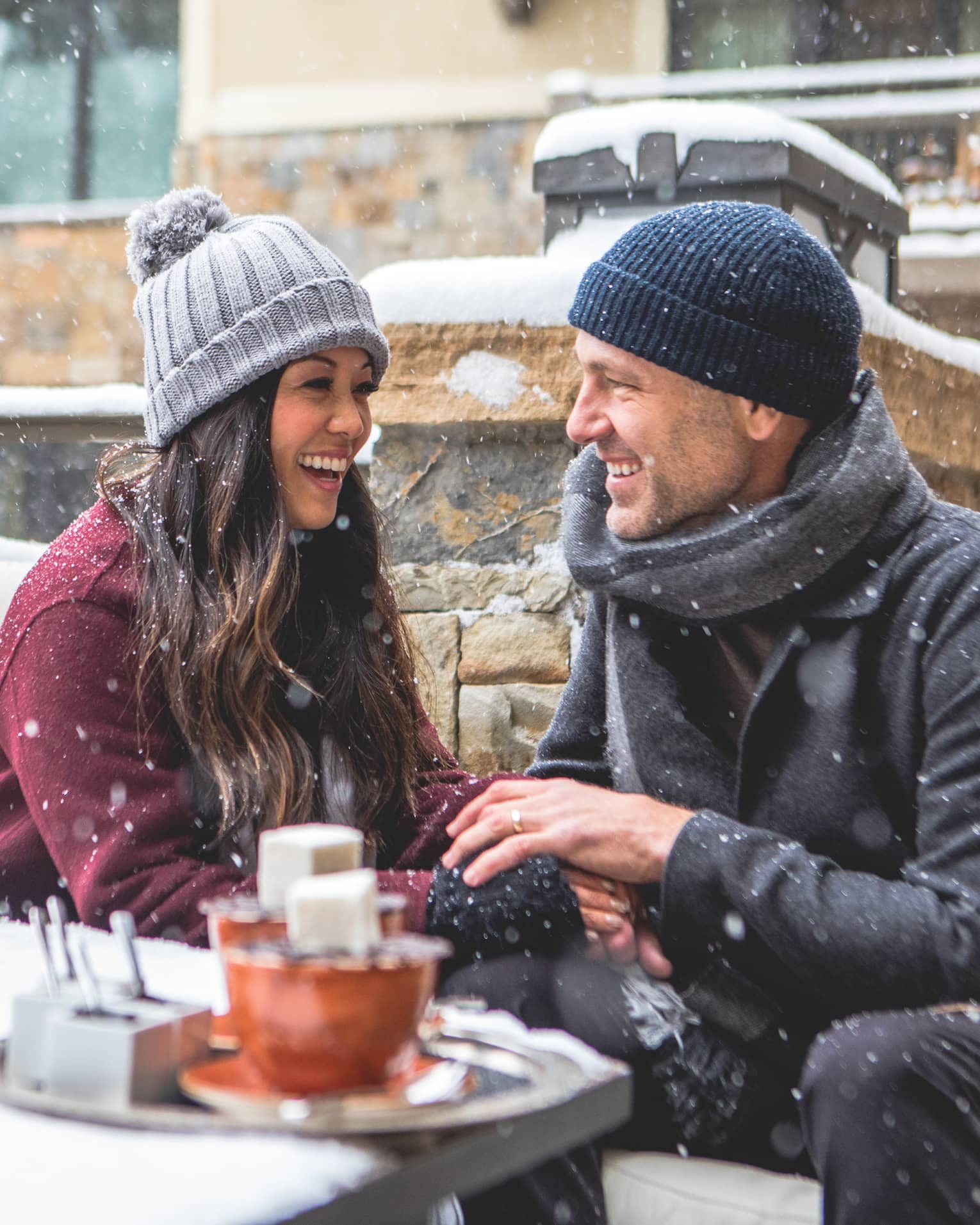 Smiling couple sitting by a firepit in a snowy outdoor setting, bundled in winter coats and hats. Snowflakes fall around them as they enjoy a cosy moment. 