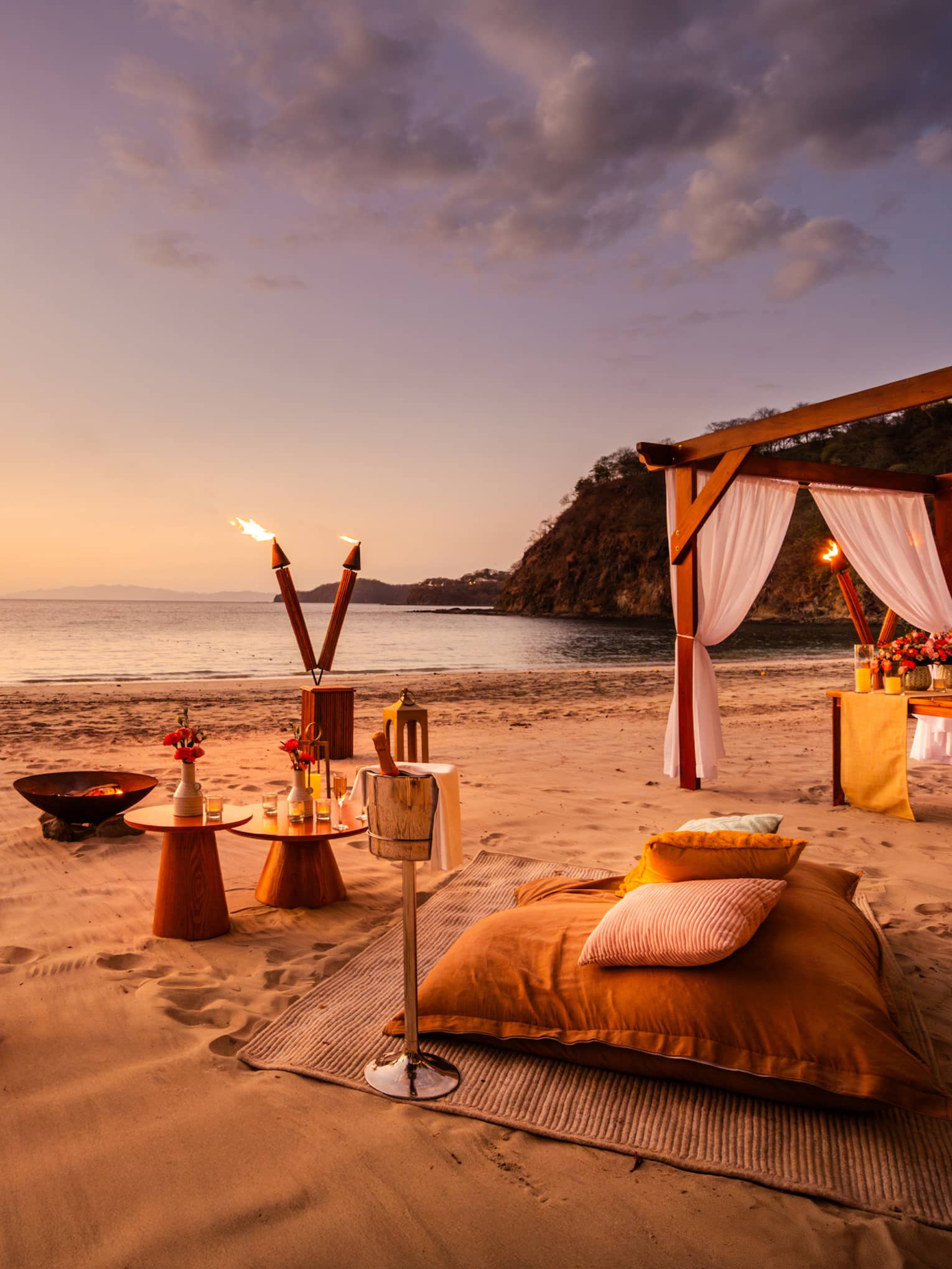 Sunset beach picnic set up with torches, lanterns, large cushions and small round tables