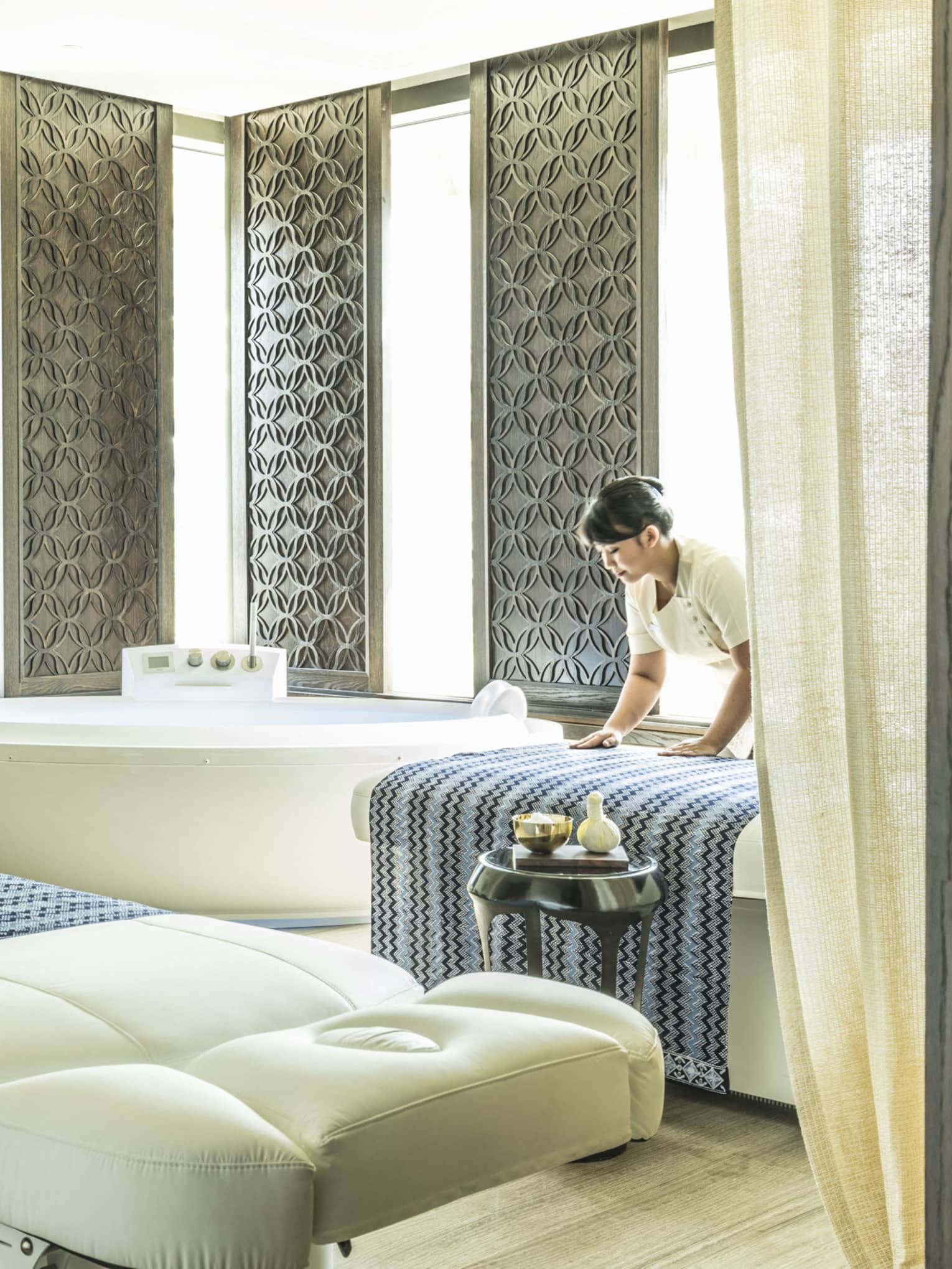Large white spa table in front of white spa tub, silver wall panels, woman places blanket on table