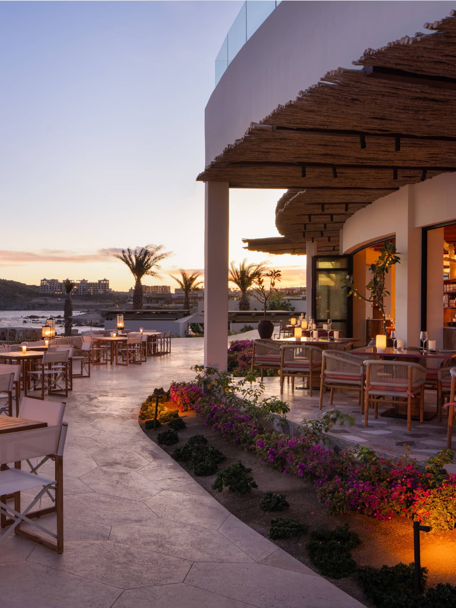 In the warm afterglow of sunset, lanterns light up a large, curved stone restaurant patio at the edge of the ocean.