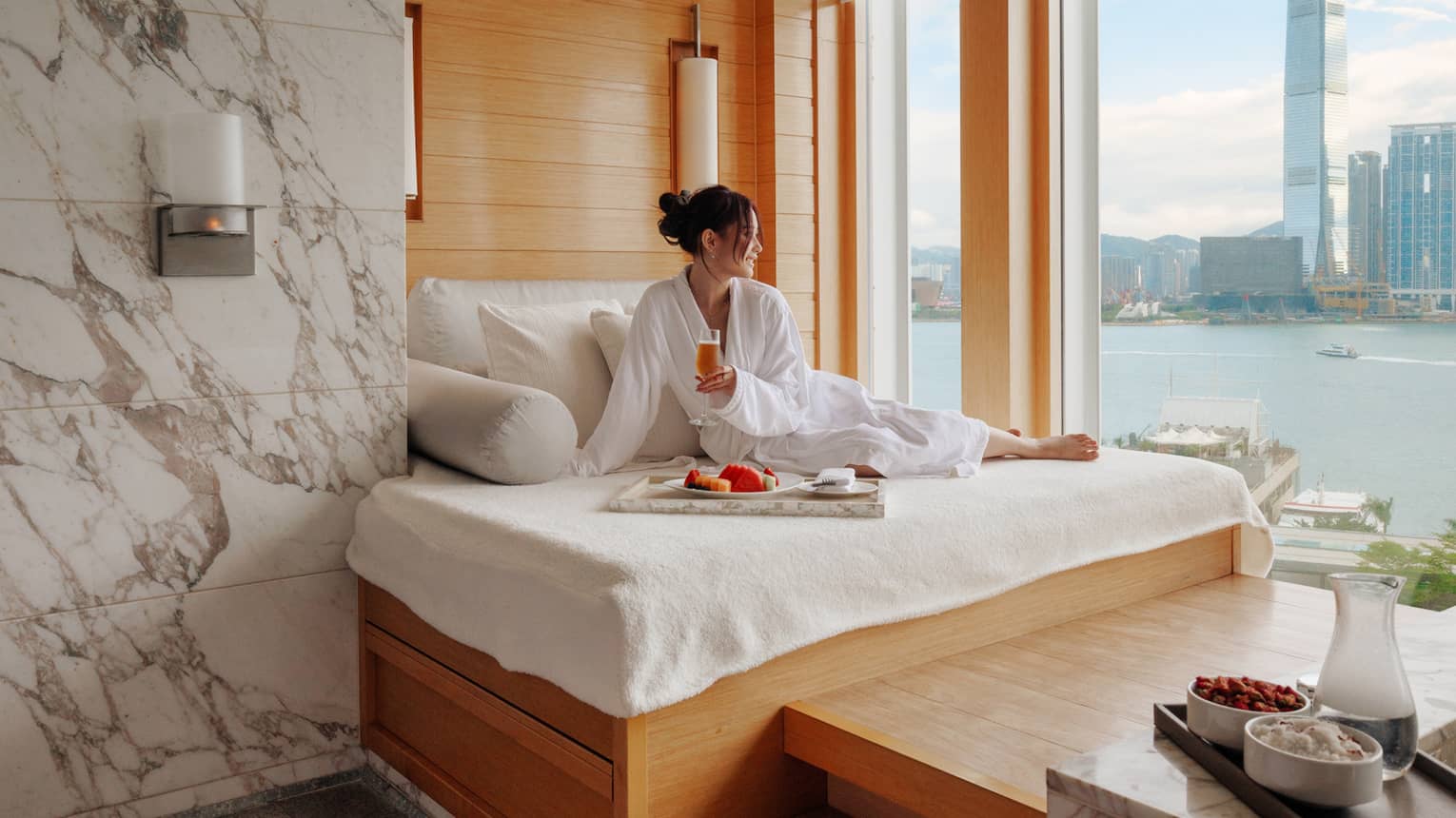 Woman in spa bathrobe sits on windowside chaise looking out to Victoria Harbour at Four Seasons Hotel Hong Kong