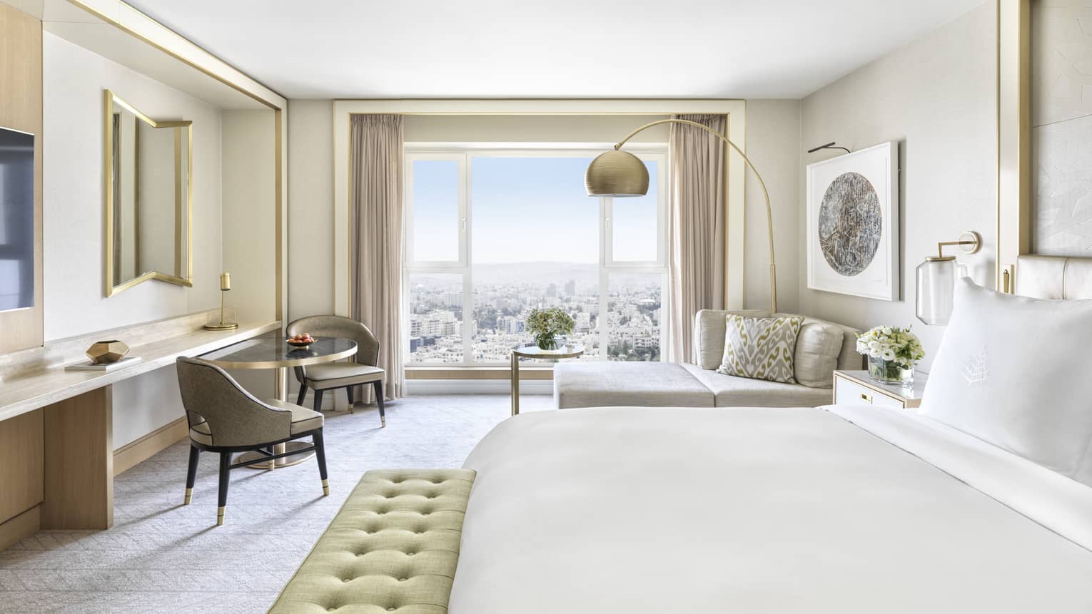 Hotel room with white king bed, light green cushioned bench, two taupe chairs, chaise, wall of windows