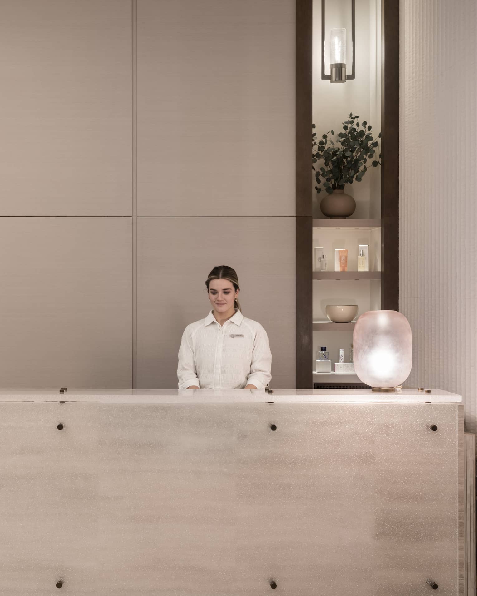 Receptionist in white shirt stands behind white marble spa reception desk with shelving display in backdrop