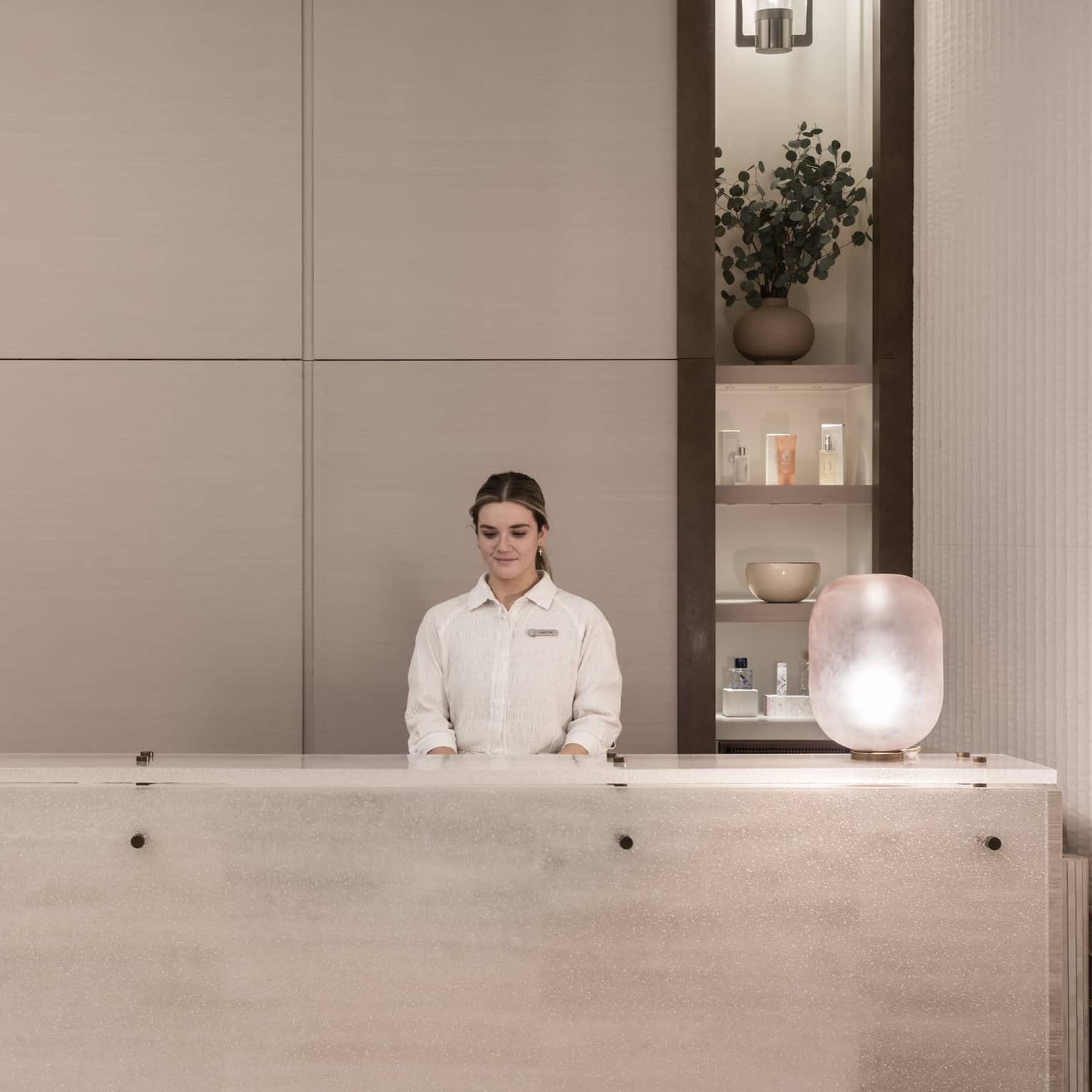 Receptionist in white shirt stands behind white marble spa reception desk with shelving display in backdrop