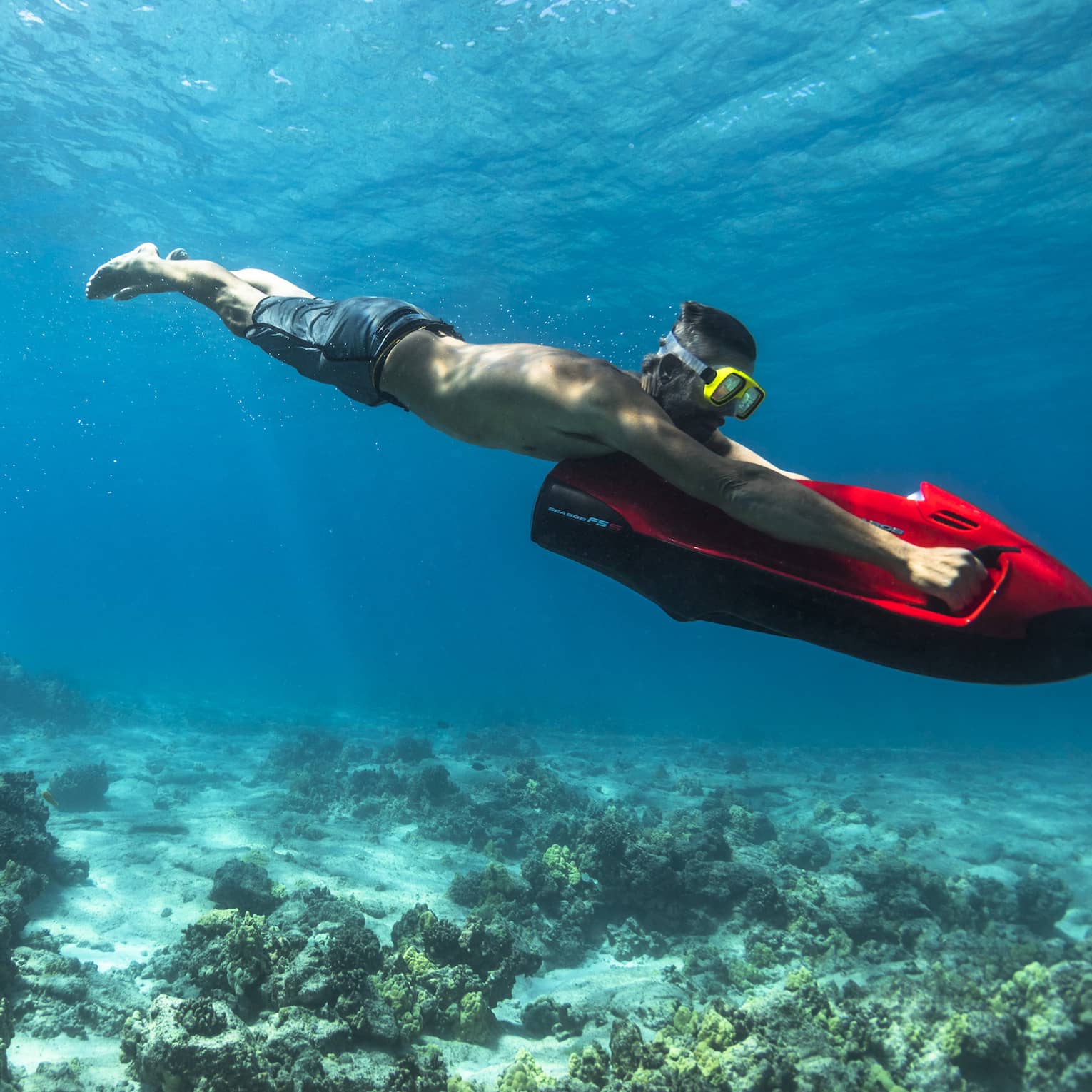 Man dives in ocean over coral, holding red inflatable board