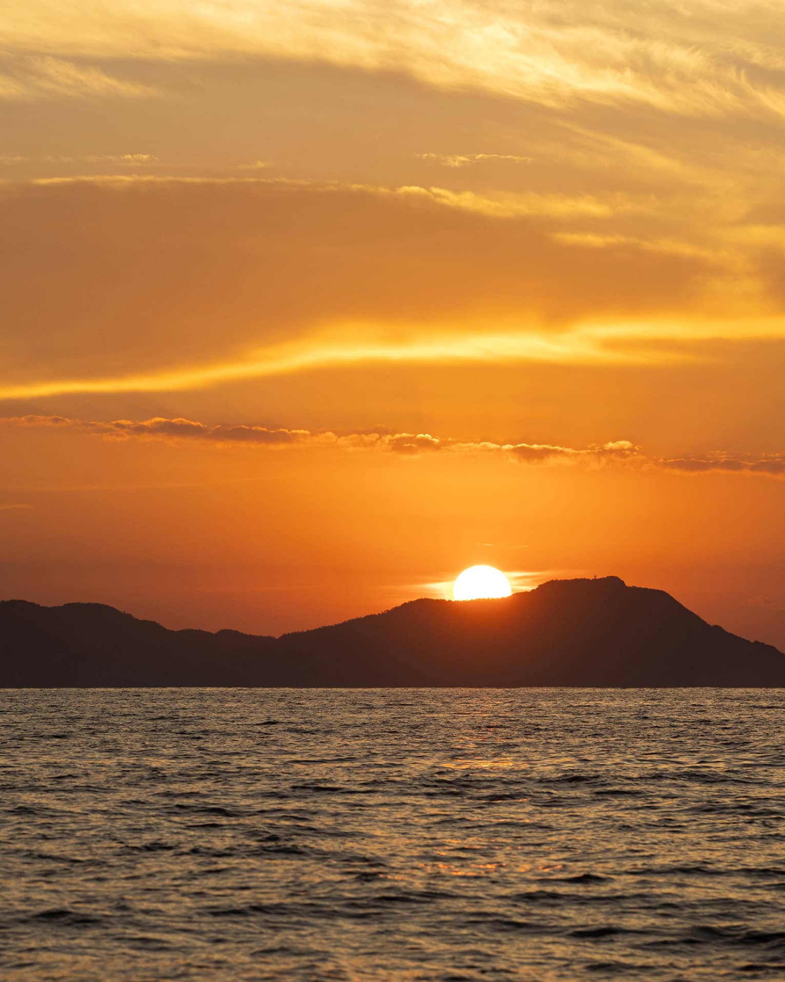 Beyond gently rippling waves, the blazing sun sets behind a hilly island, wispy clouds in the sky awash in its orange glow.