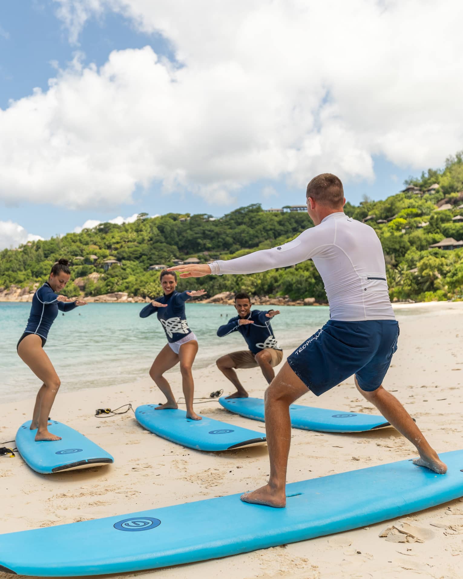 Surfing instructor balances on surfboard on white sand beach in front of row of surfers
