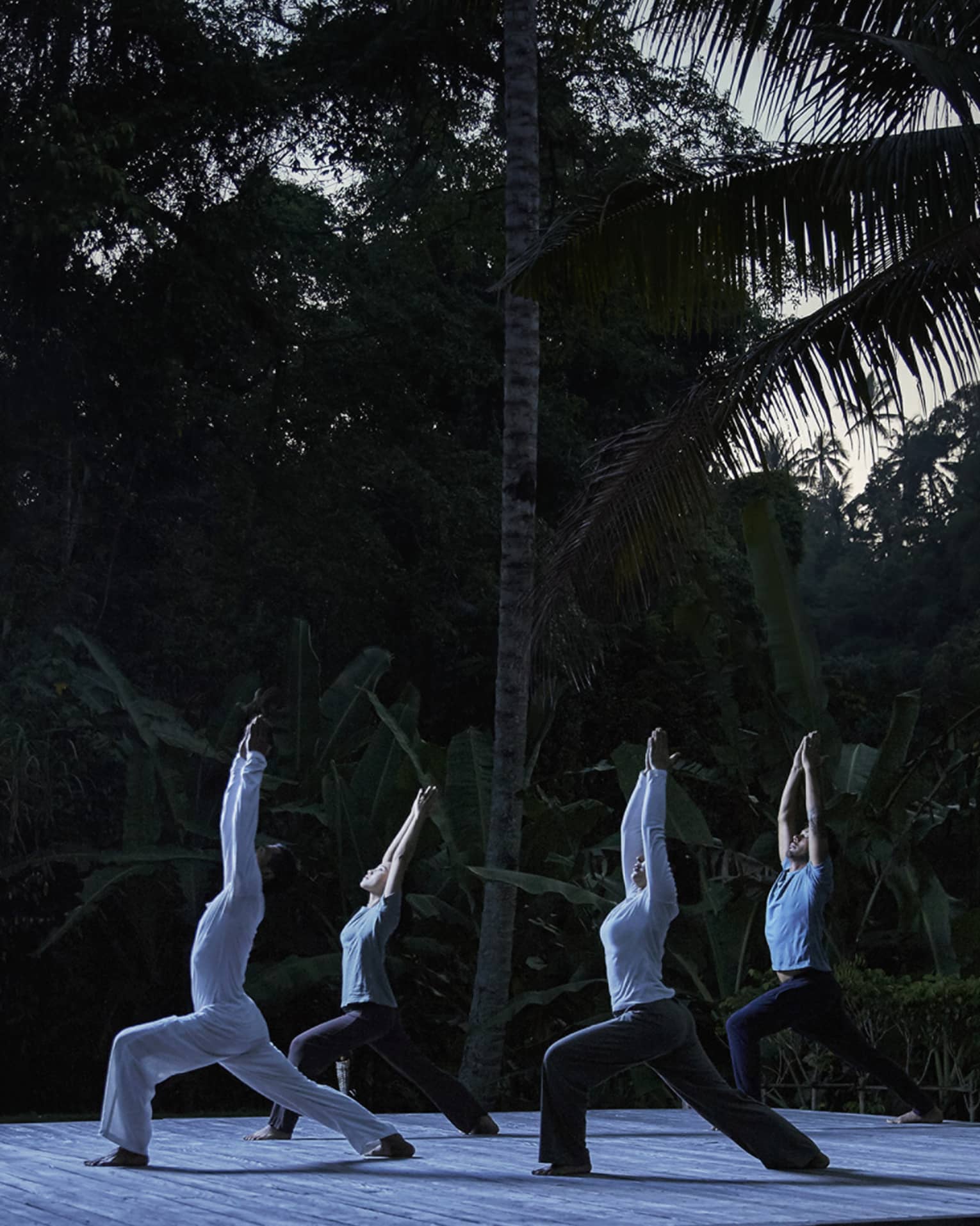 Five people kneel in yoga pose with arms above head on wood platform in forest at night