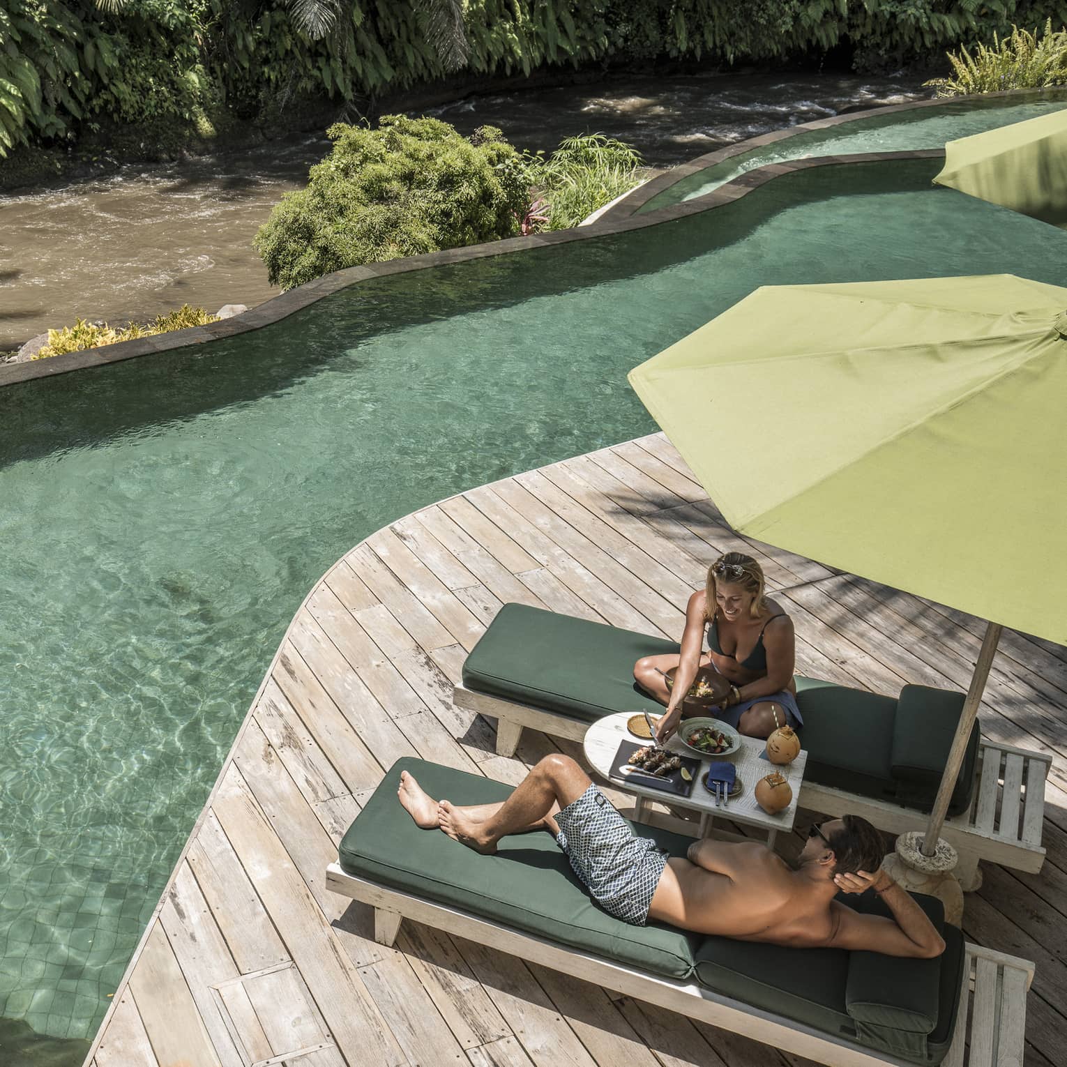 Couple lounges, dines on patio chairs on deck by outdoor swimming pool
