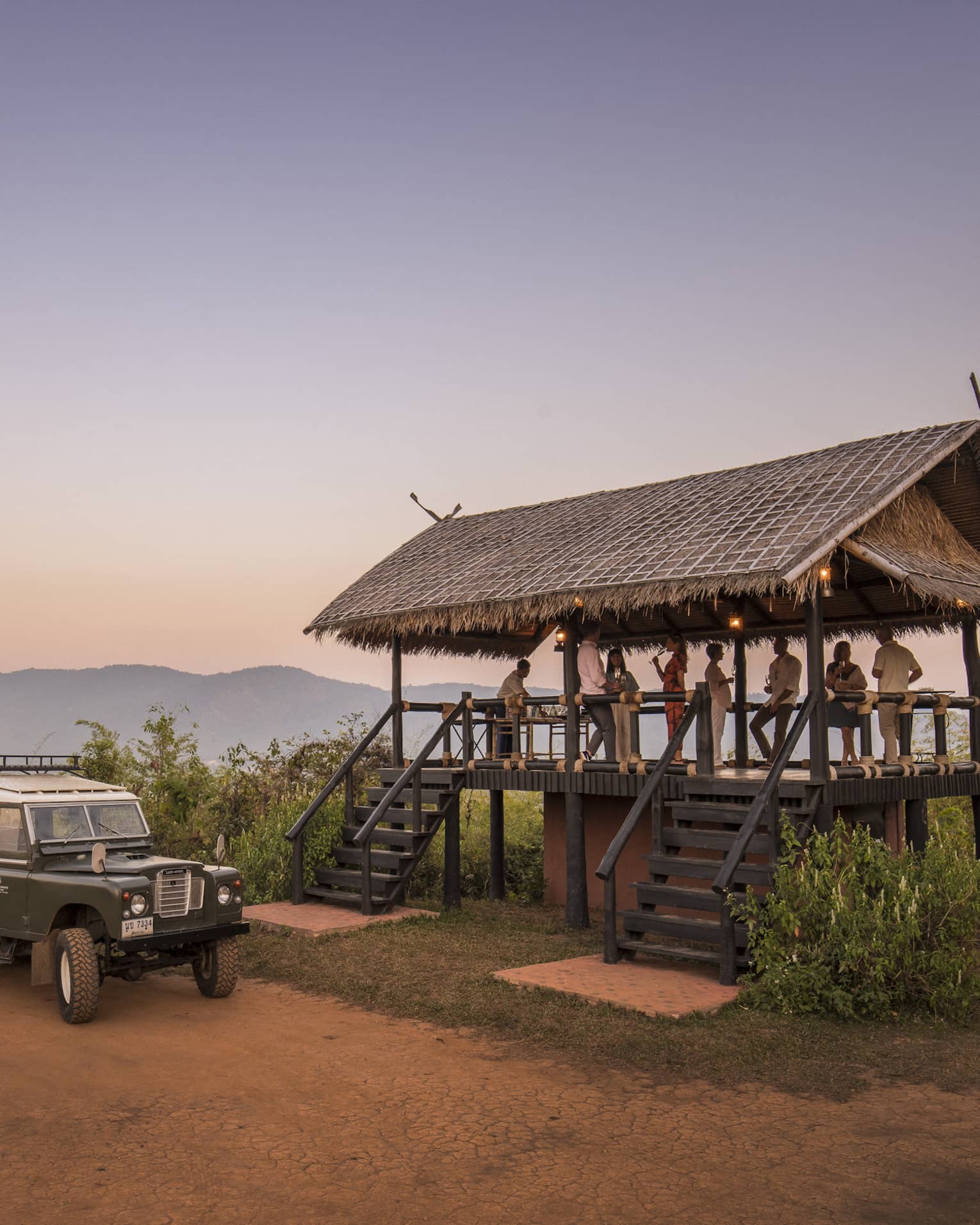 People gather on a raised thatched deck near a campfire and off-road vehicle at sunset.