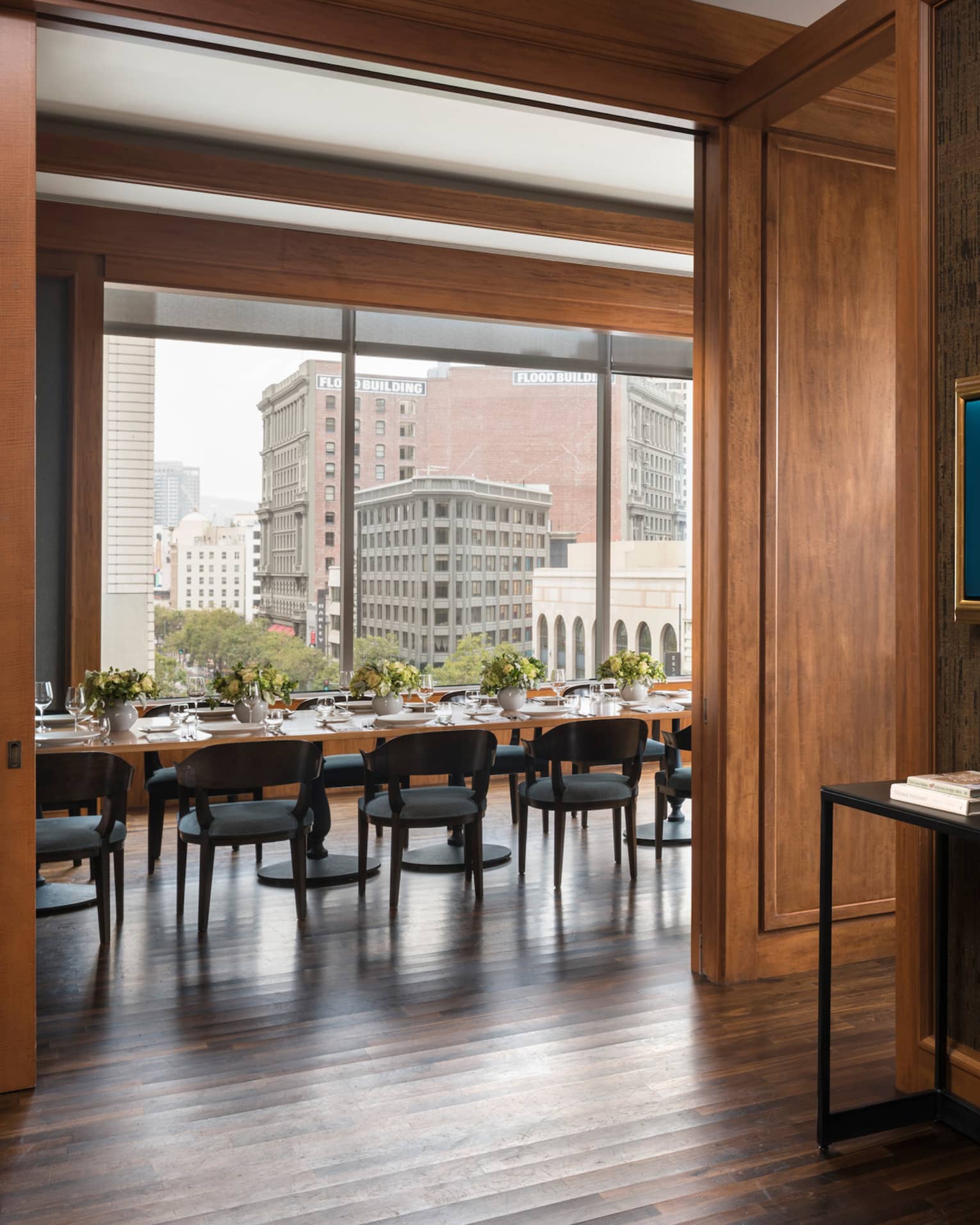 A waiting area in a restaurant with grey chairs and a small table, a private dining area with large windows can be seen through a hall.