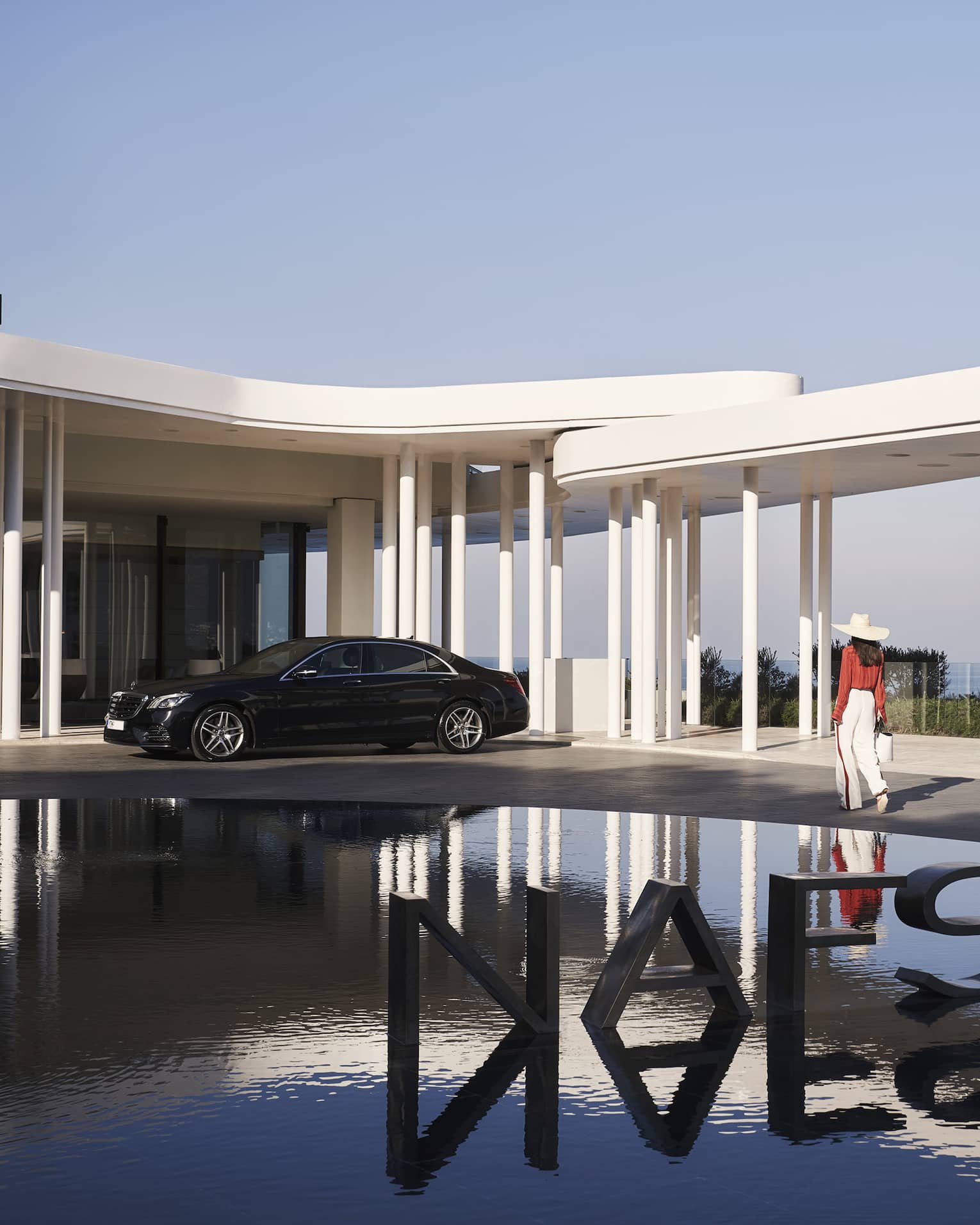 A guest passes a metal sign reading NAFSIKA in a fountain and towards an elegant car parked under modern white porticos.