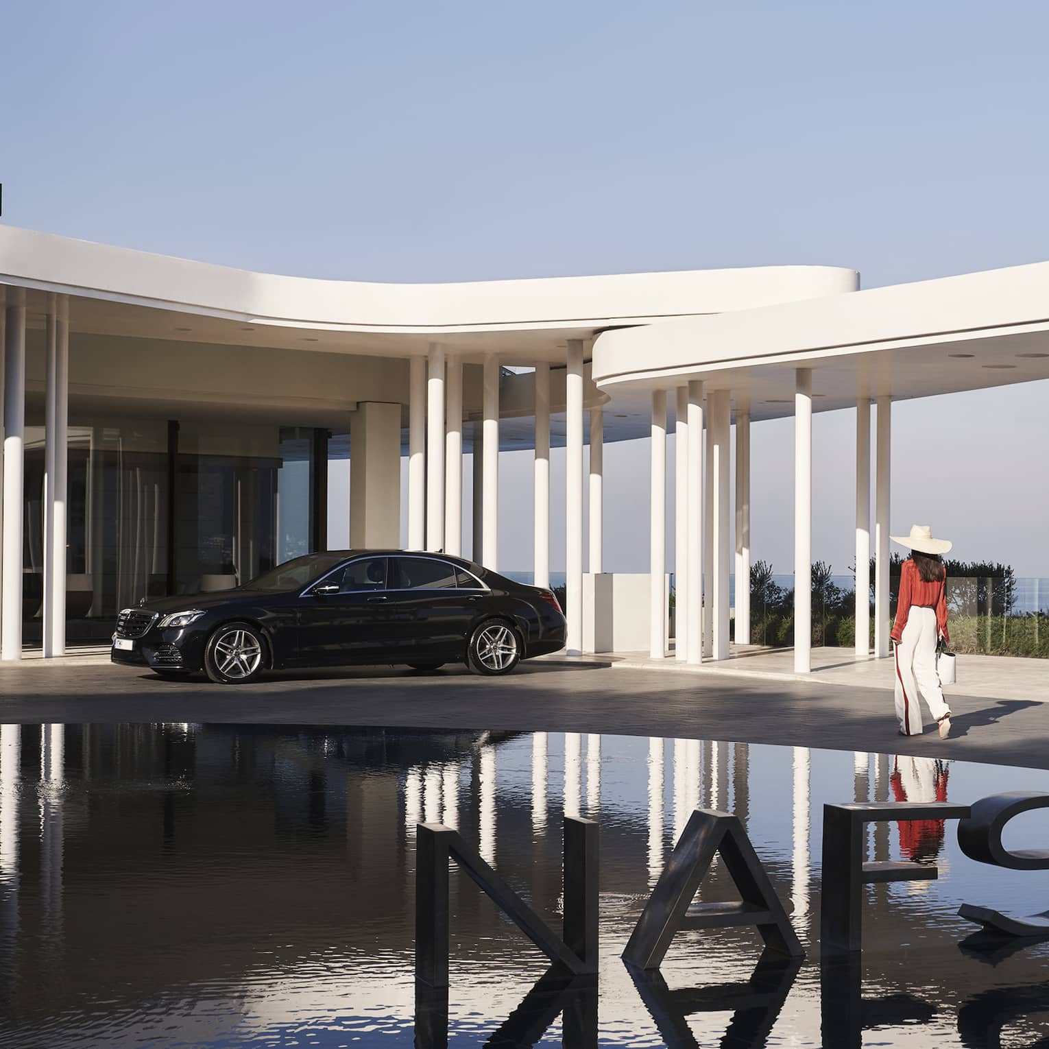 A guest passes a metal sign reading NAFSIKA in a fountain and towards an elegant car parked under modern white porticos.