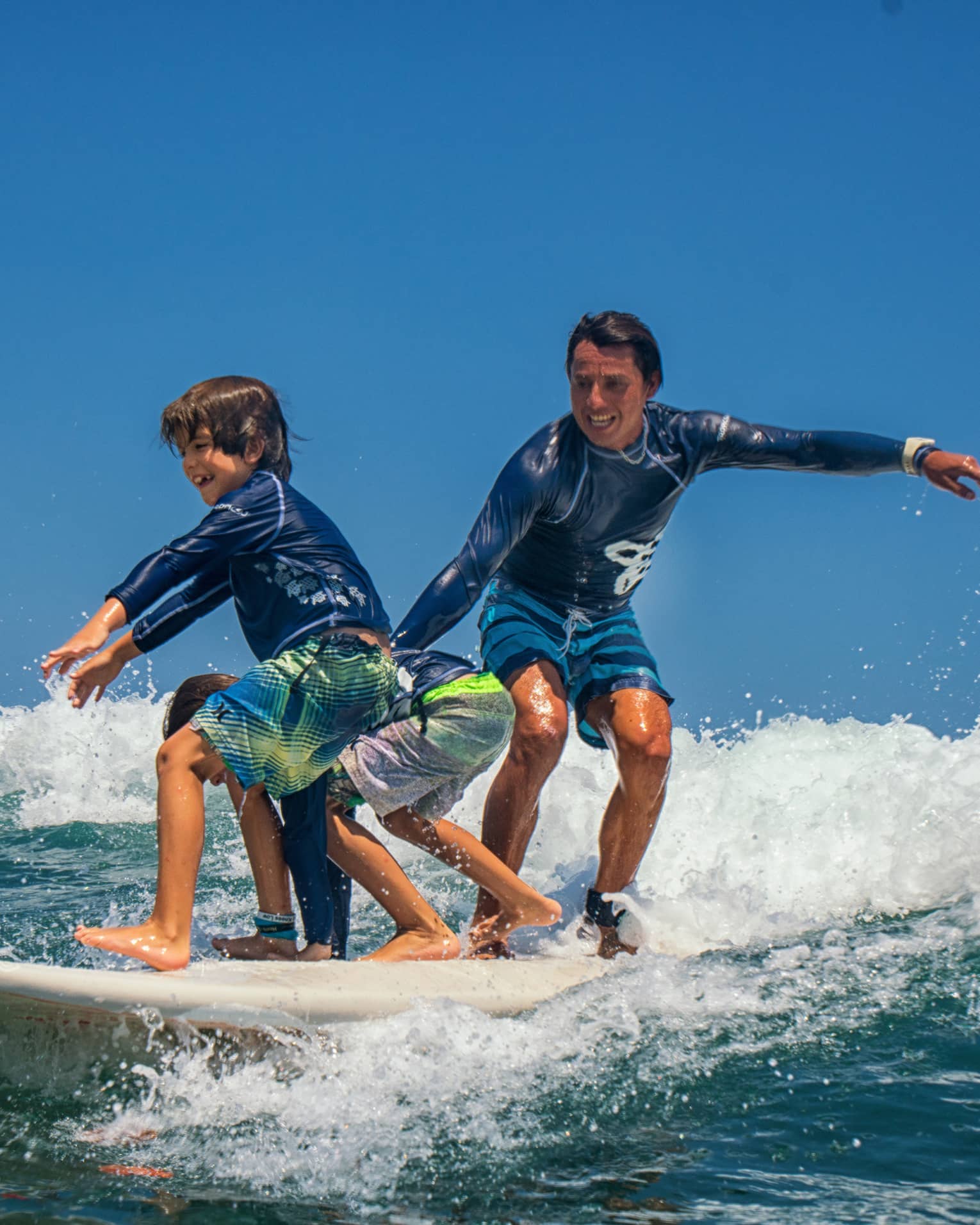 Adult and two children share a surfboard, laughing as they balance on a foamy wave. 