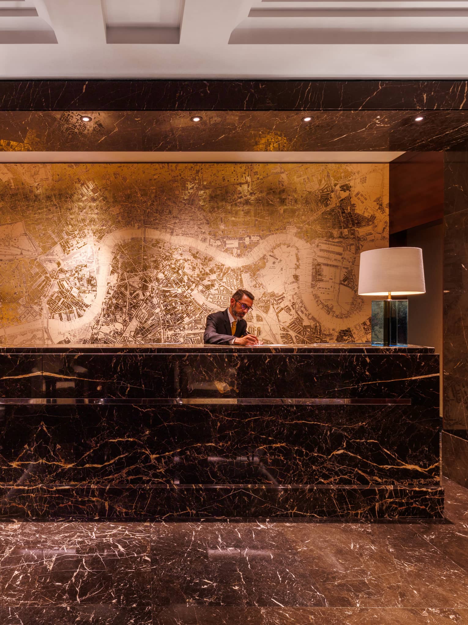 A warm, softly lit hotel lobby space with a person behind a large desk, wall decor, and both desktop and ceiling lighting
