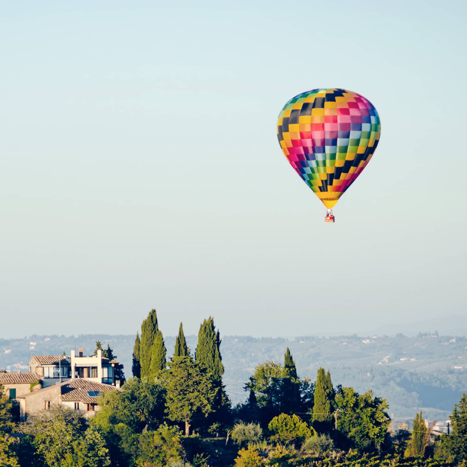 Colourful hot air balloon floating above a landscape with trees and houses, set against a clear blue sky.		