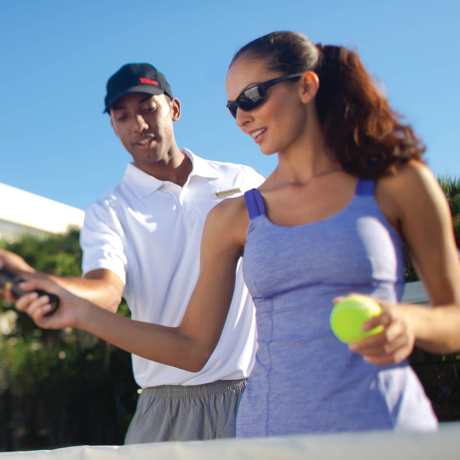 Close-up of instructor teaching a guest how to grip the racquet for a serve under clear blue sky on an outdoor tennis court.