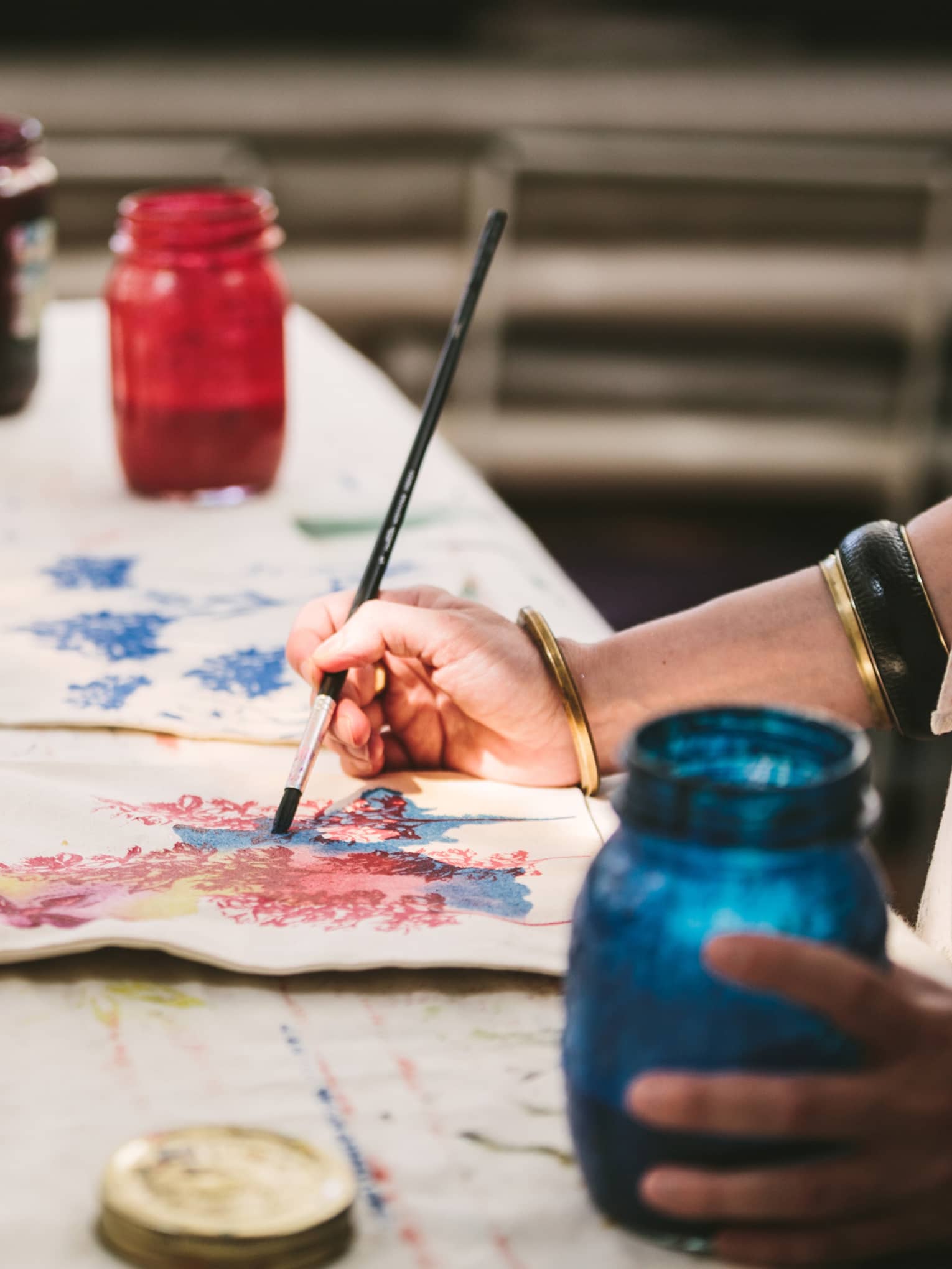 An artist using mason jars filled with red, blue, yellow and black paint to paint images of flowers on canvas tote bags.