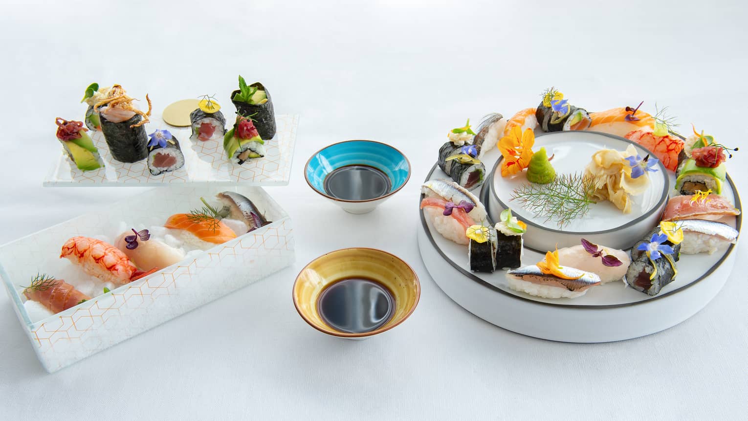 Three dishes of assorted sushi with two bowls of dipping sauce between