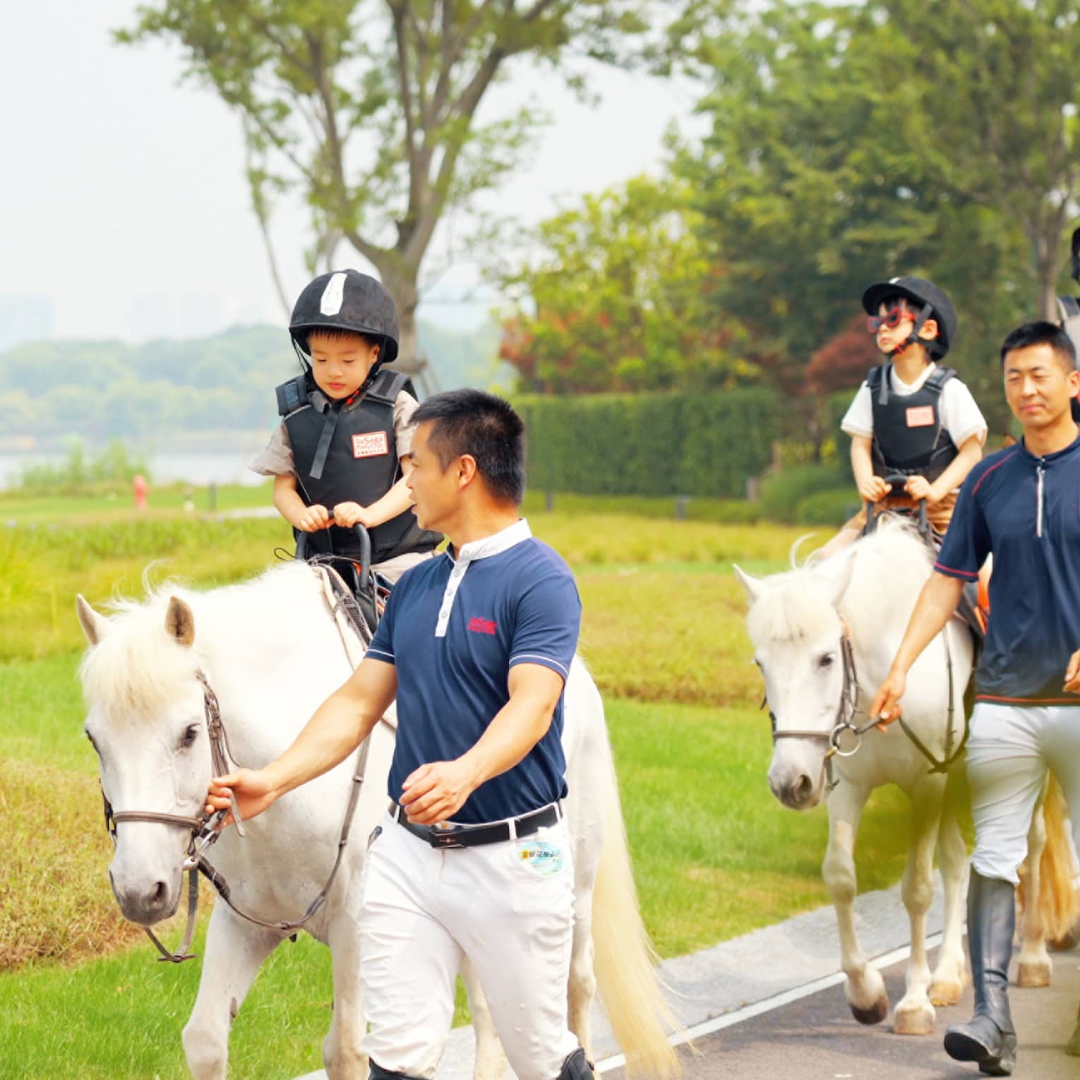 Three small children and an adult in riding gear walk approach atop white ponies, four guides gently holding their bridles.