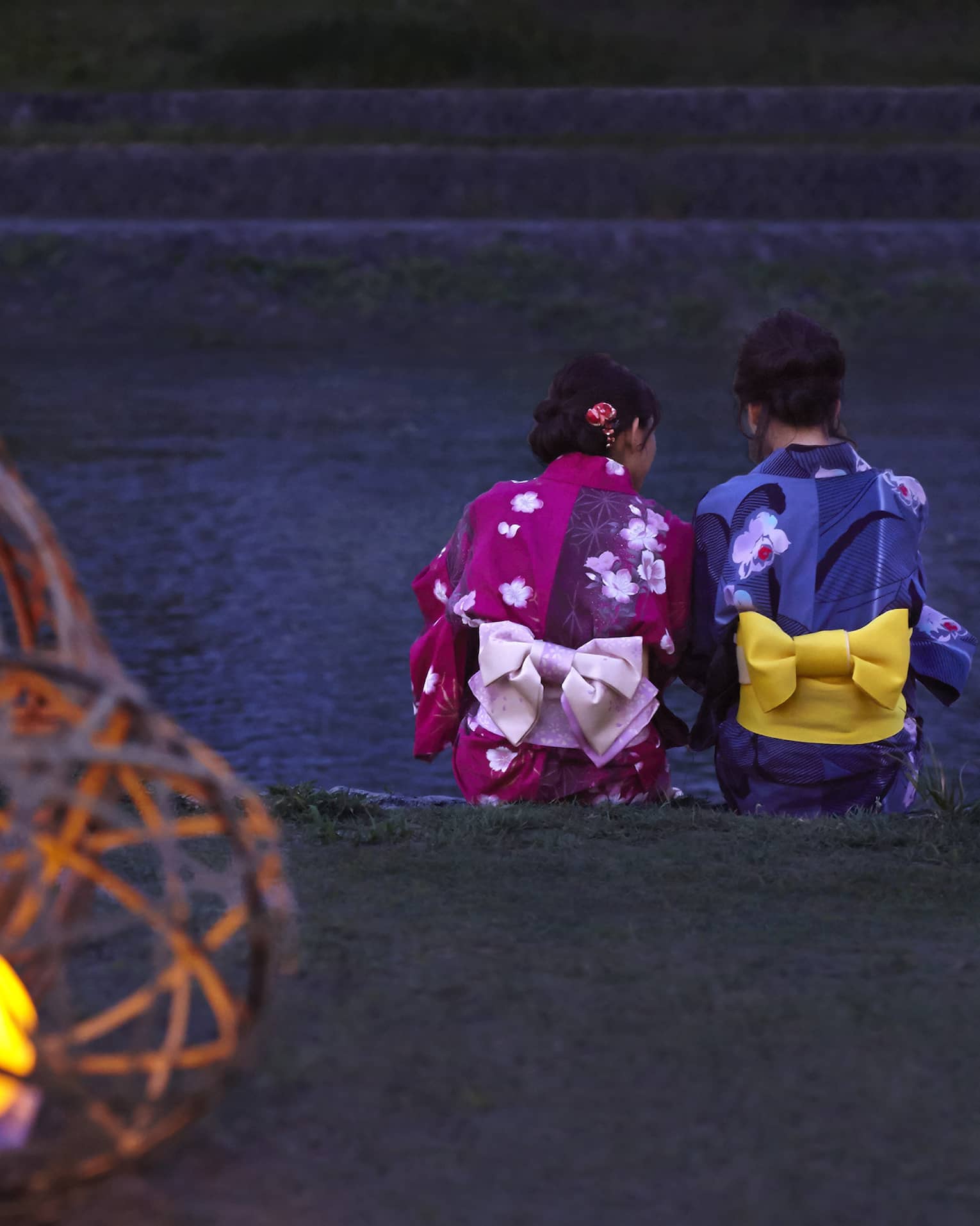 Two Geisha sit on ledge by a river at dusk