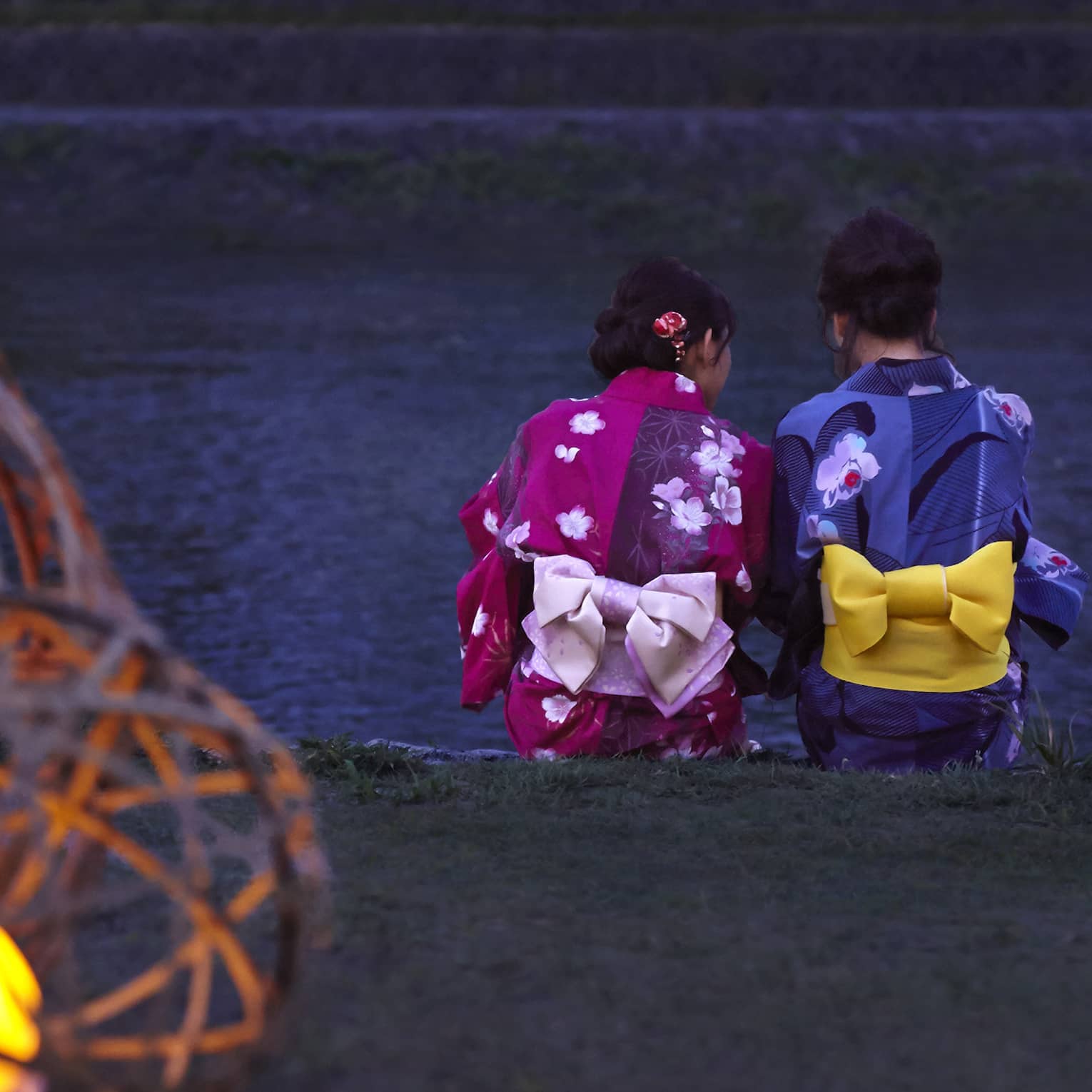 Two Geisha sit on ledge by a river at dusk
