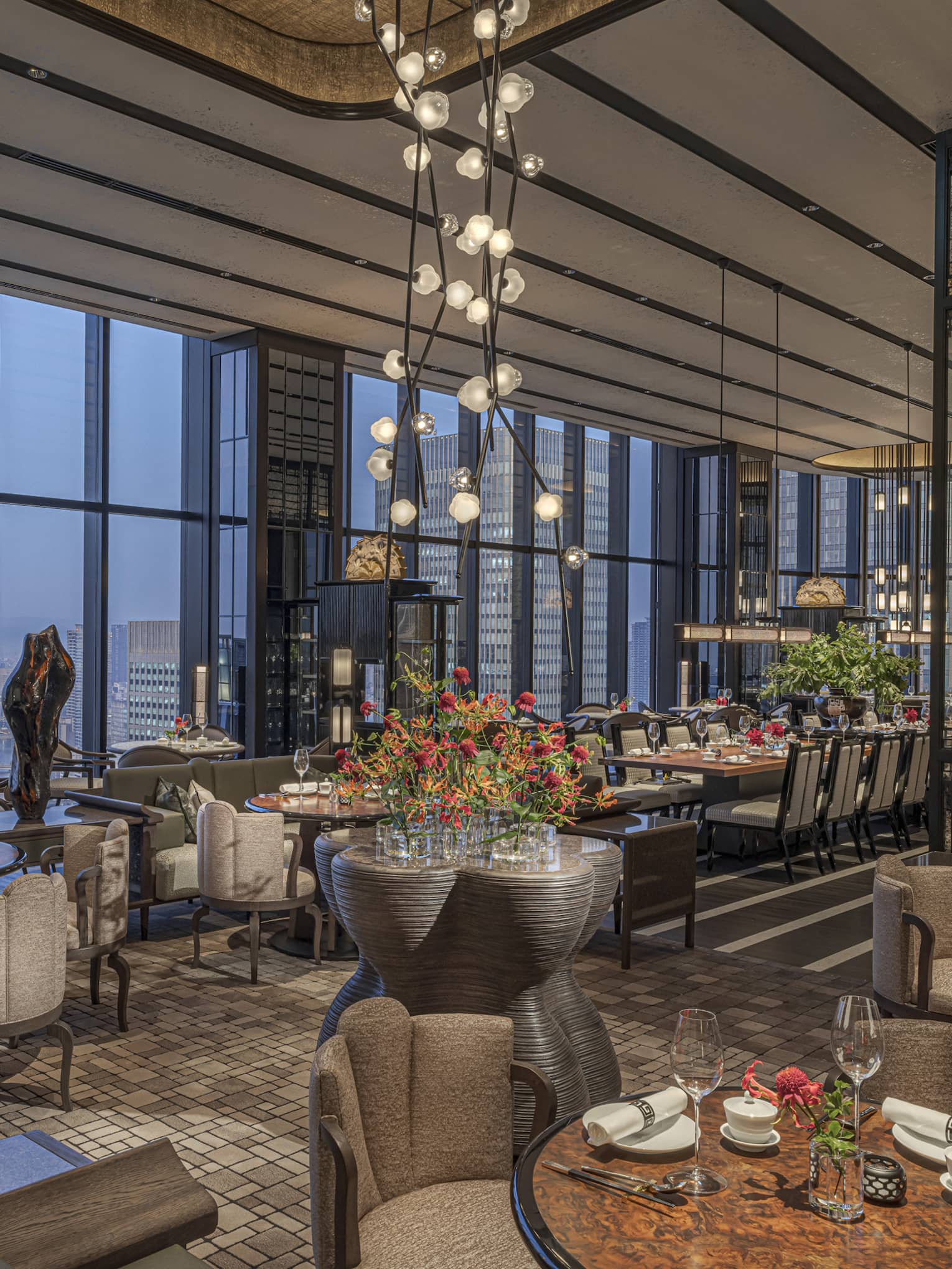 Expansive high-end restaurant warmly lit by modern chandeliers with a bank of windows offering aerial views of the city