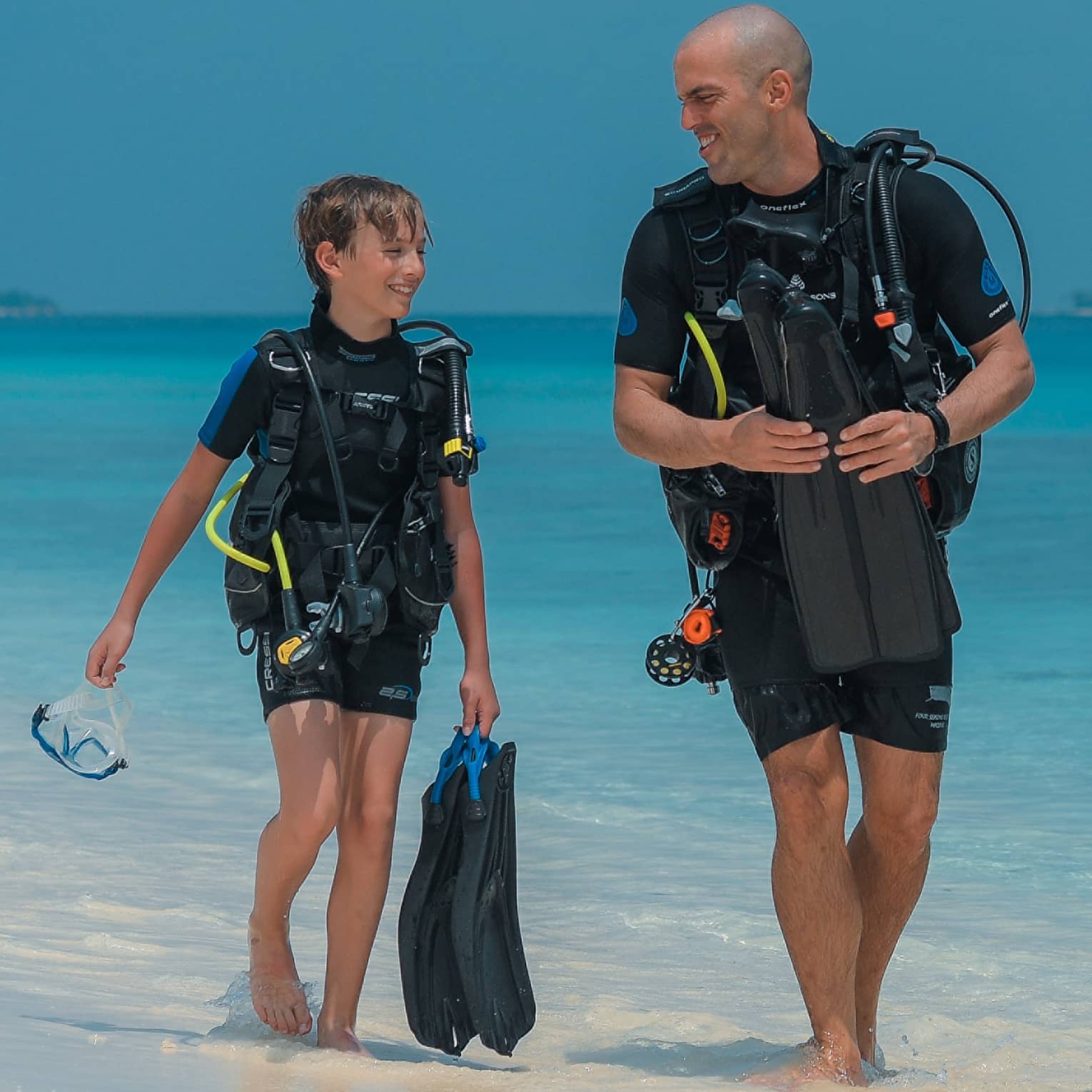Smiling child and scuba instructor in wetsuits and scuba gear walk at water's edge, aqua ocean behind, cloudless sky above.