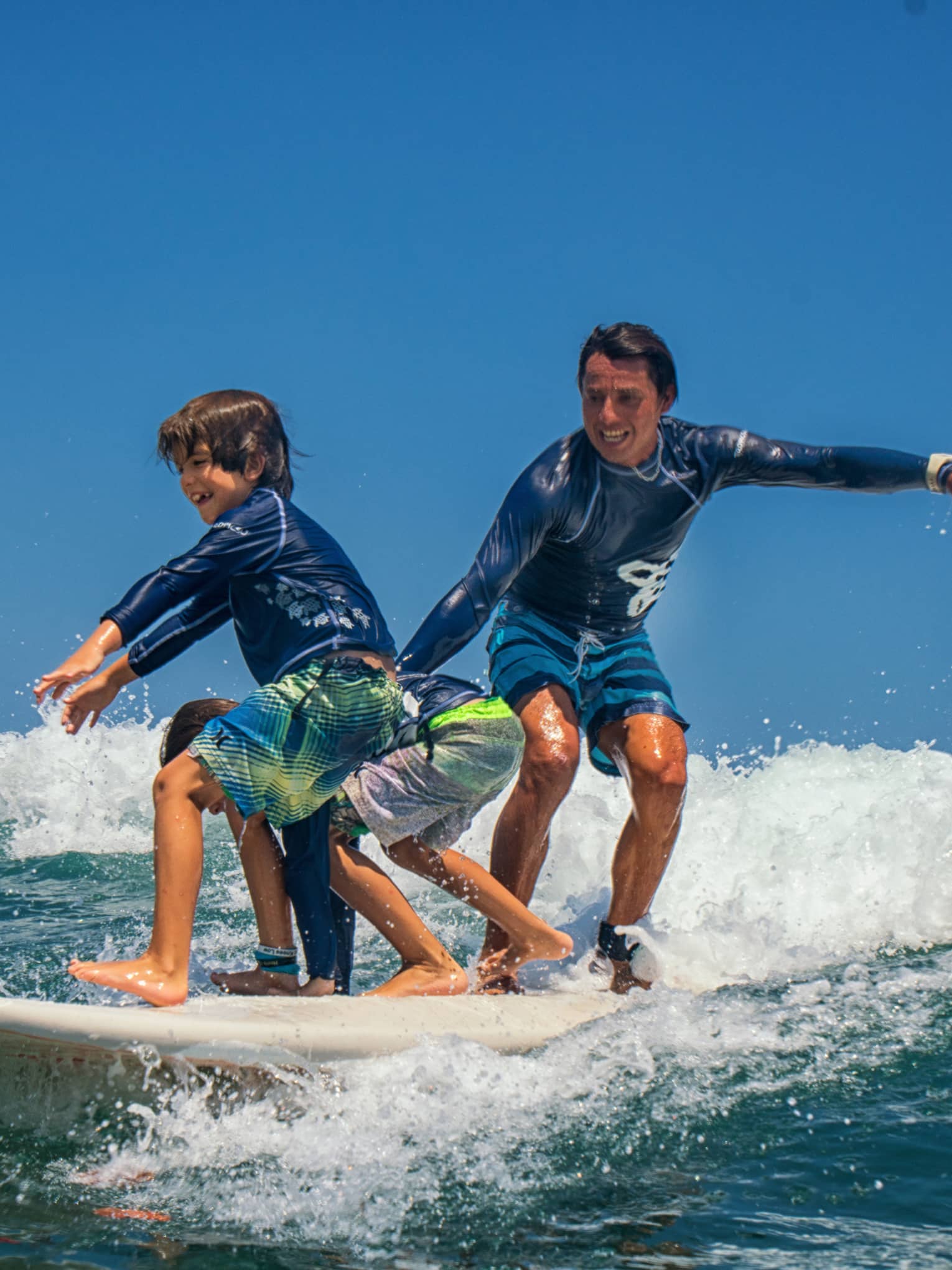 Adult and two children share a surfboard, laughing as they balance on a foamy wave. 