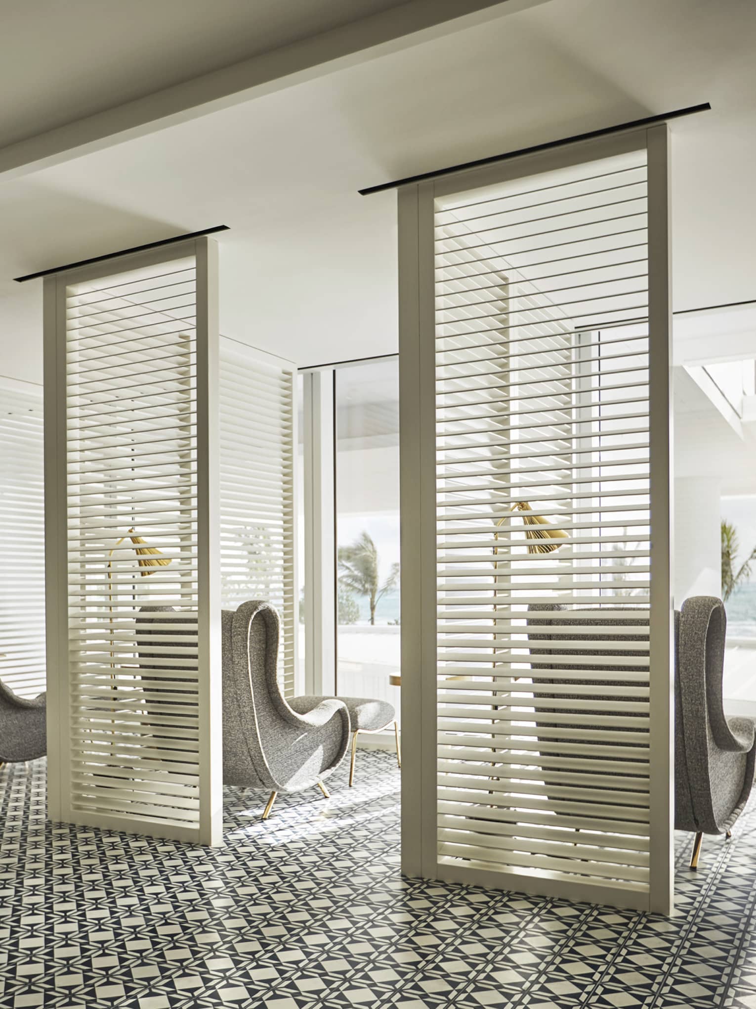 Luxury spa area with patterned tile flooring, partitioned seating space, gray armchairs and floor-to-ceiling windows overlooking ocean and palm trees