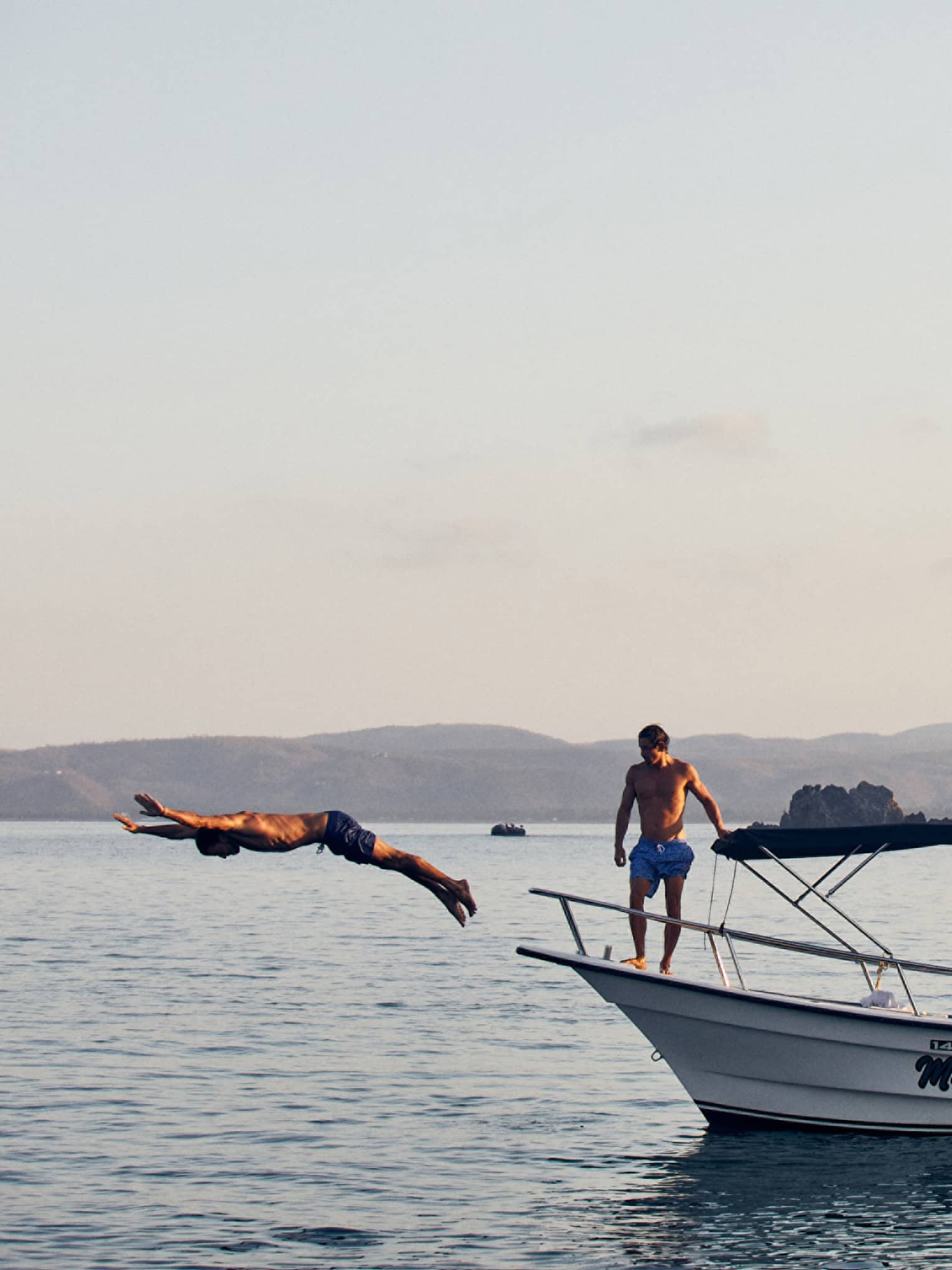 Side view of a guest diving gracefully into the water while others watch from a boat's deck, distant landmasses beyond.