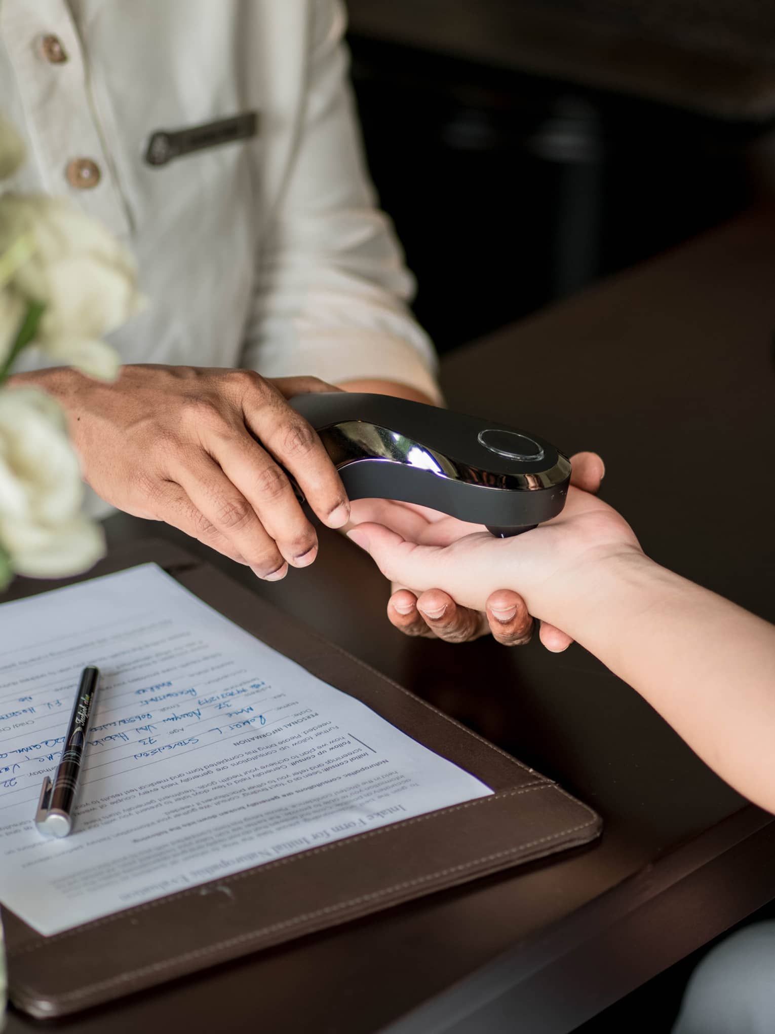 A person scans another's palm with a smooth, shiny, black electronic device beside a completed medical form on the table..